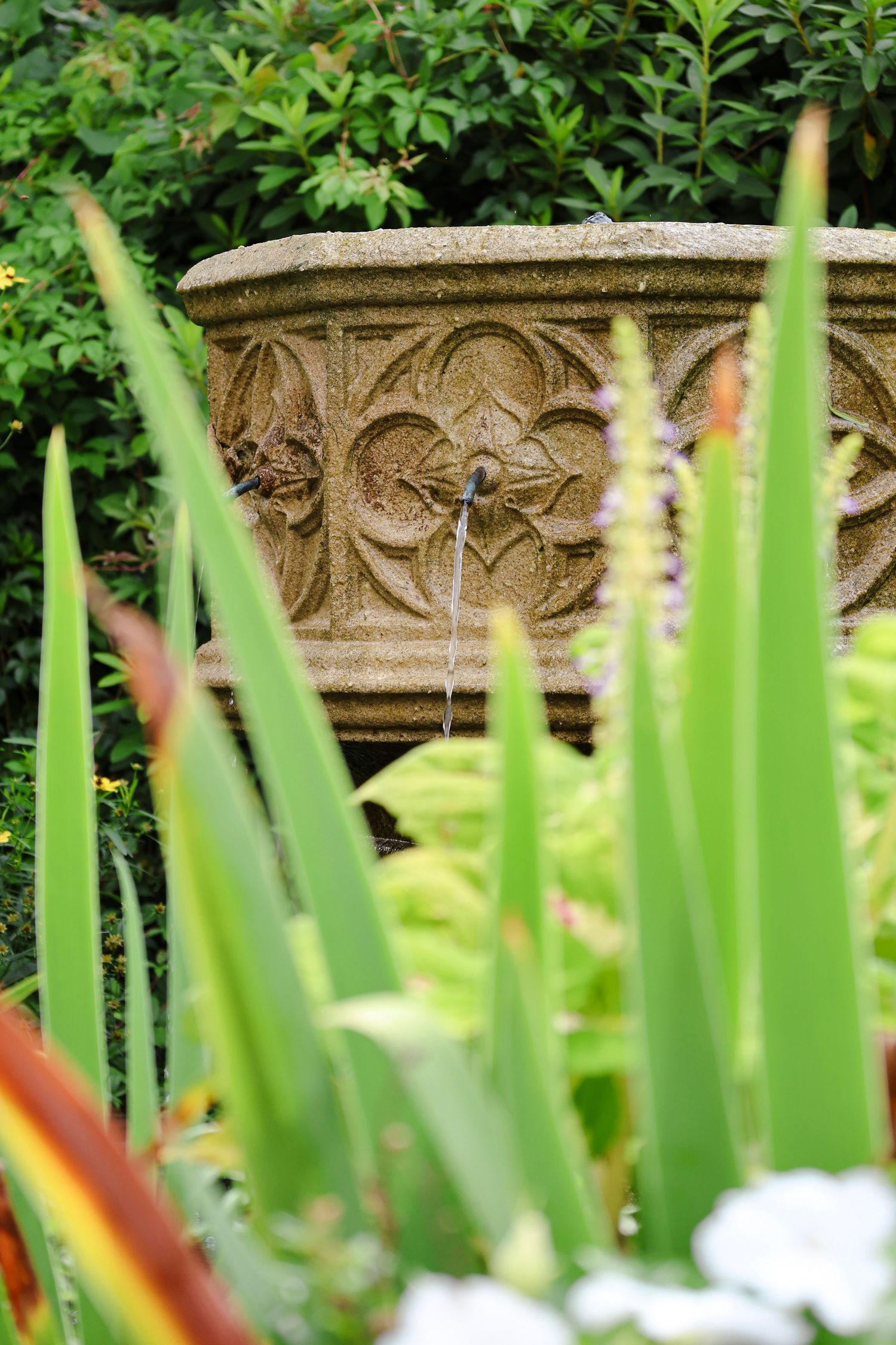 Stone garden fountain with ornate carvings surrounded by lush green plants and colorful flowers in a serene garden setting.