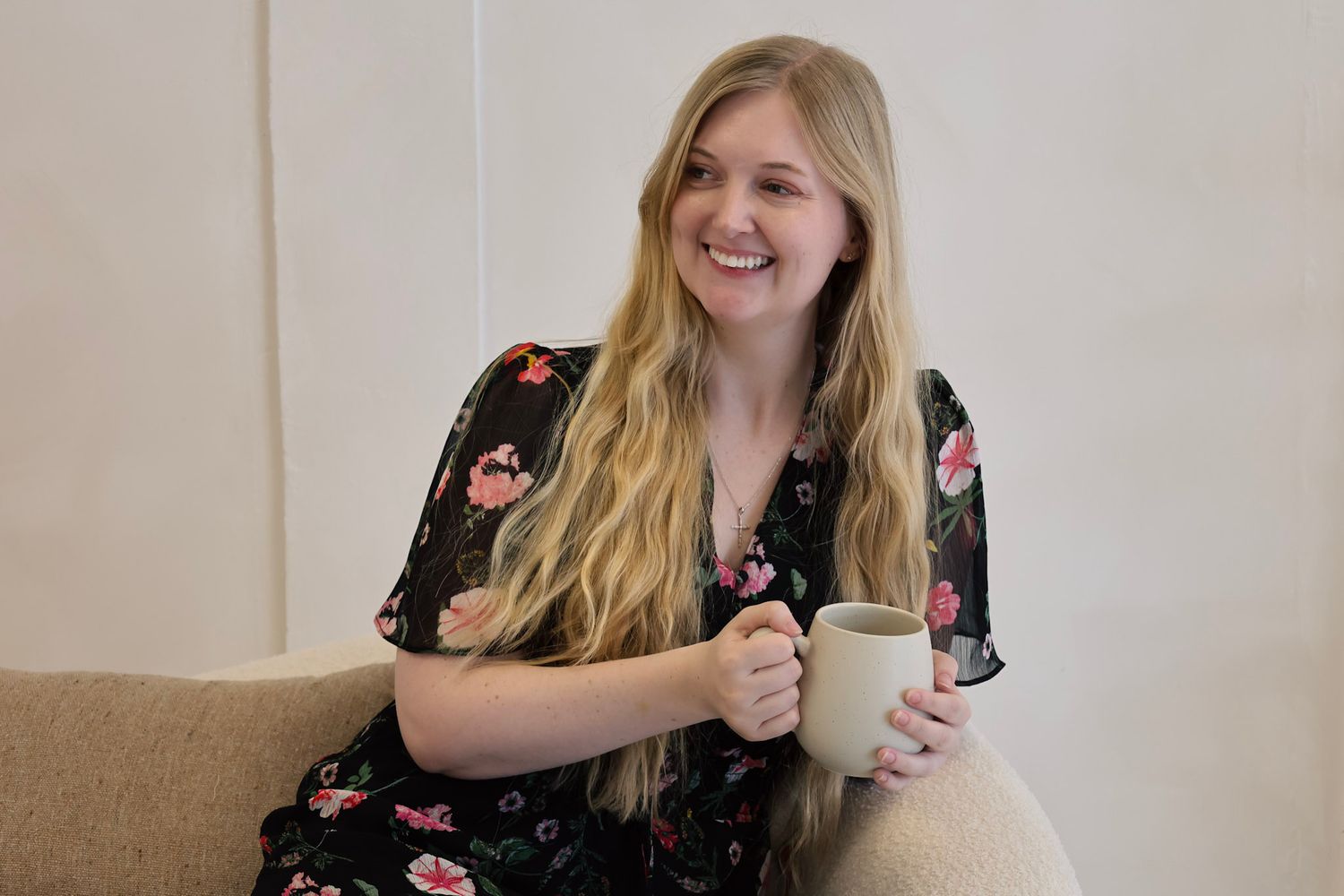 Smiling young blonde woman in floral dress holding a mug, sitting on a couch in a bright modern interior.
