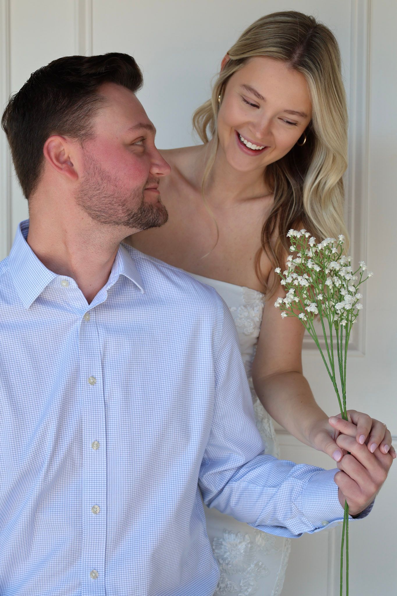 A smiling bride holding baby's breath flowers stands behind a man in a blue shirt in a romantic couple portrait.