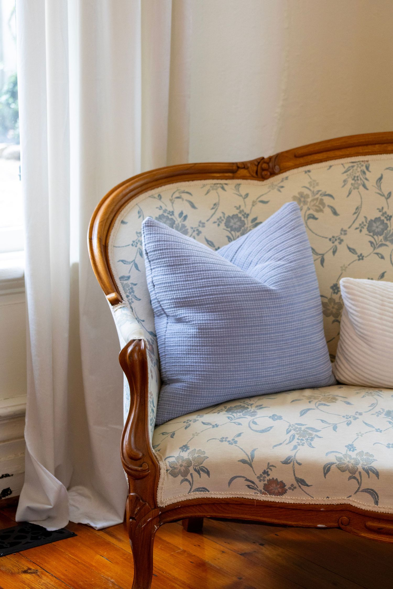 Vintage French-style loveseat with floral upholstery and wooden frame, styled with blue textured throw pillows near a window.