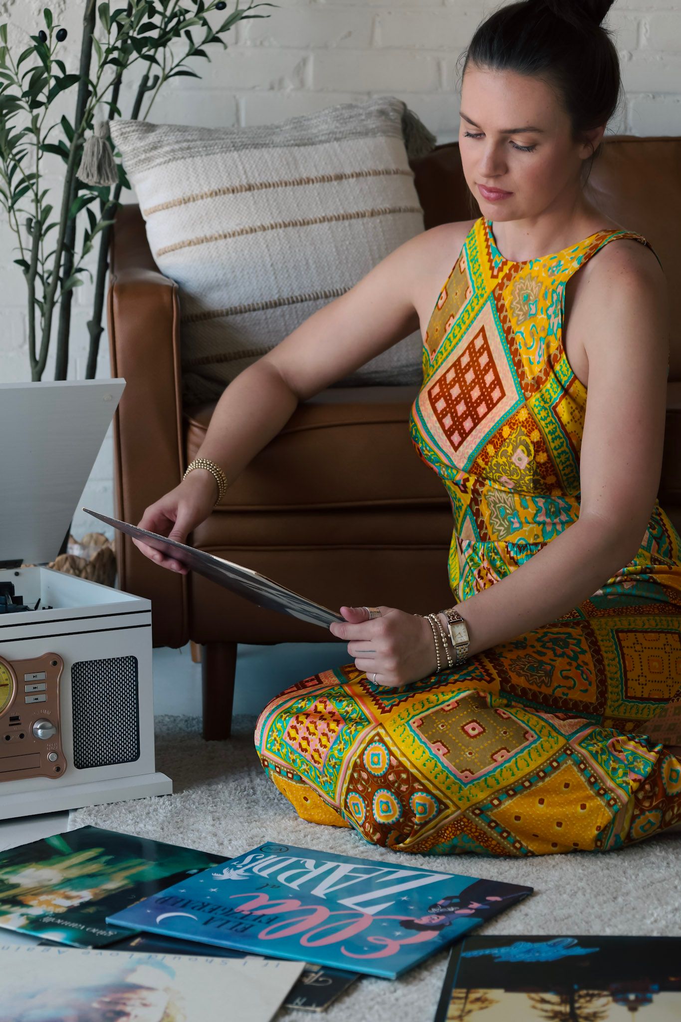 Woman in colorful patterned dress sitting cross-legged on floor, browsing vinyl records near a turntable.