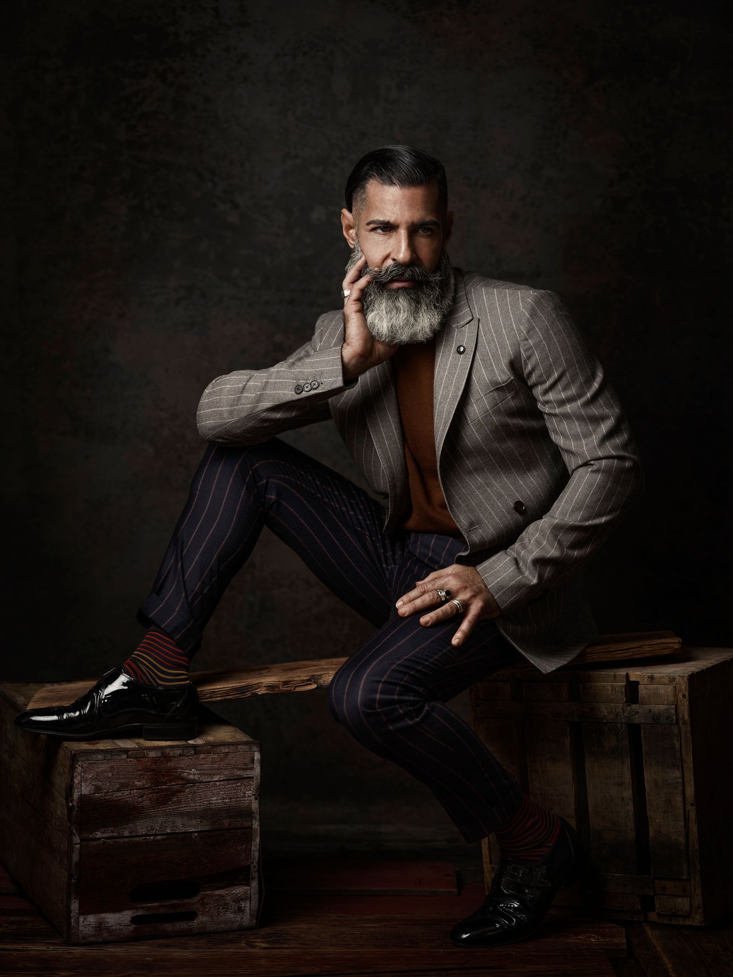 Stylish bearded man in pinstripe suit seated on wooden crate.