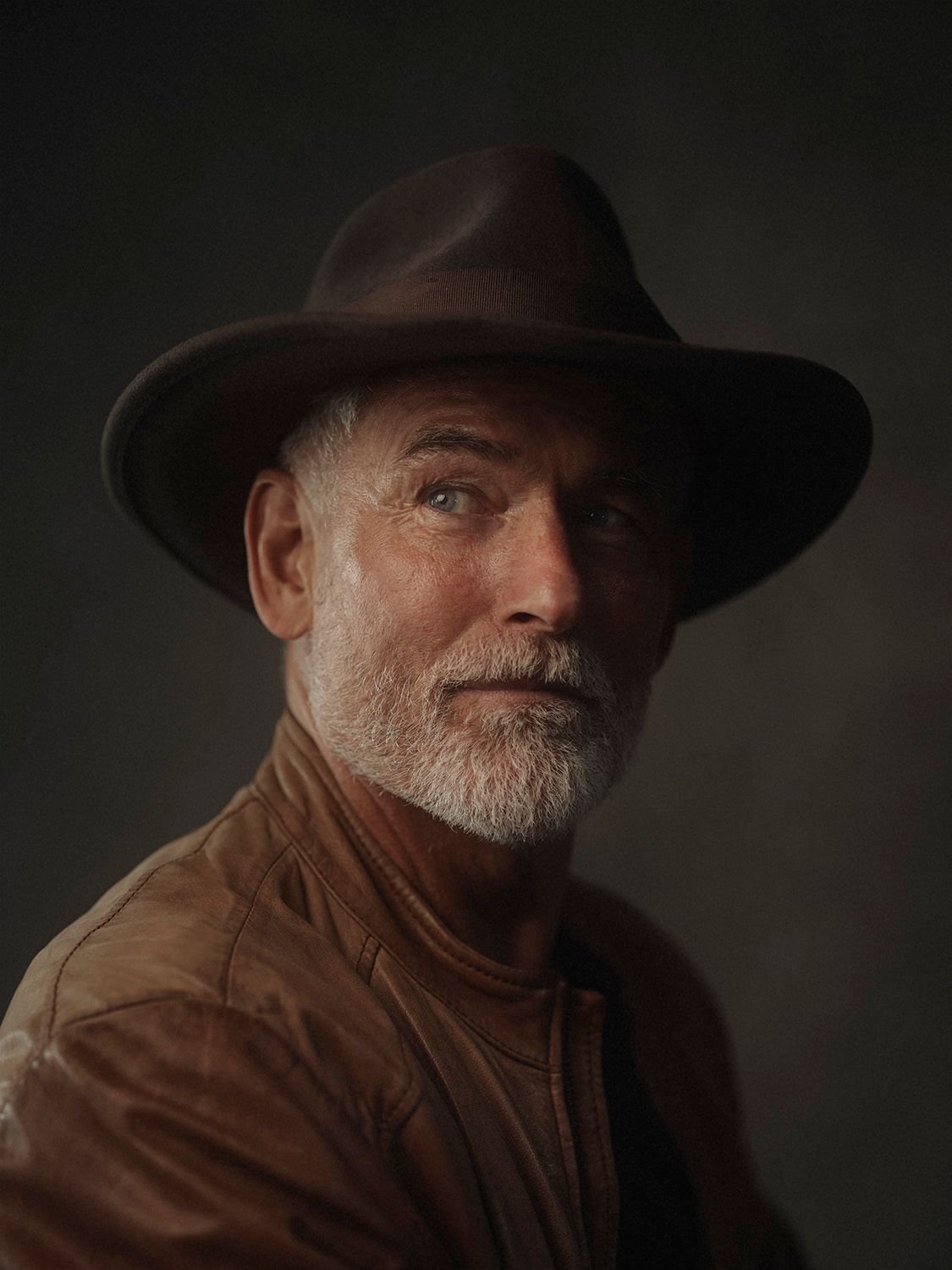 Portrait of an older man with a white beard wearing a brown leather jacket and wide-brim hat.