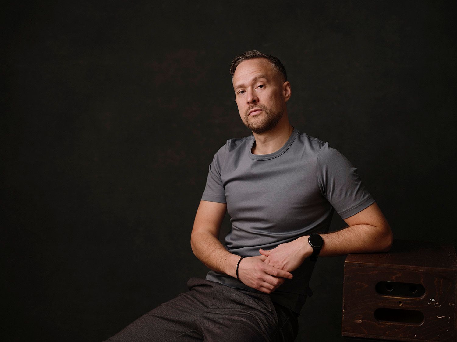 Man in gray t-shirt sitting on a wooden box against a dark background in a professional portrait.