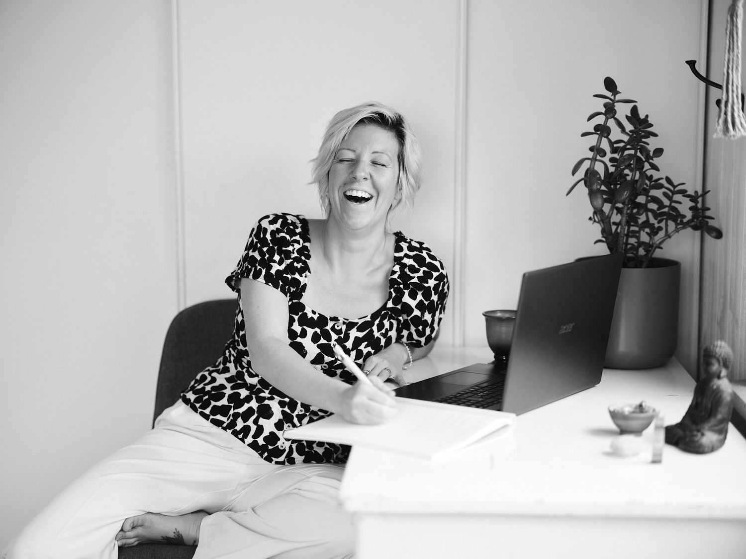 Black and white photo of a laughing blonde woman sitting at a desk with a laptop and notebook.