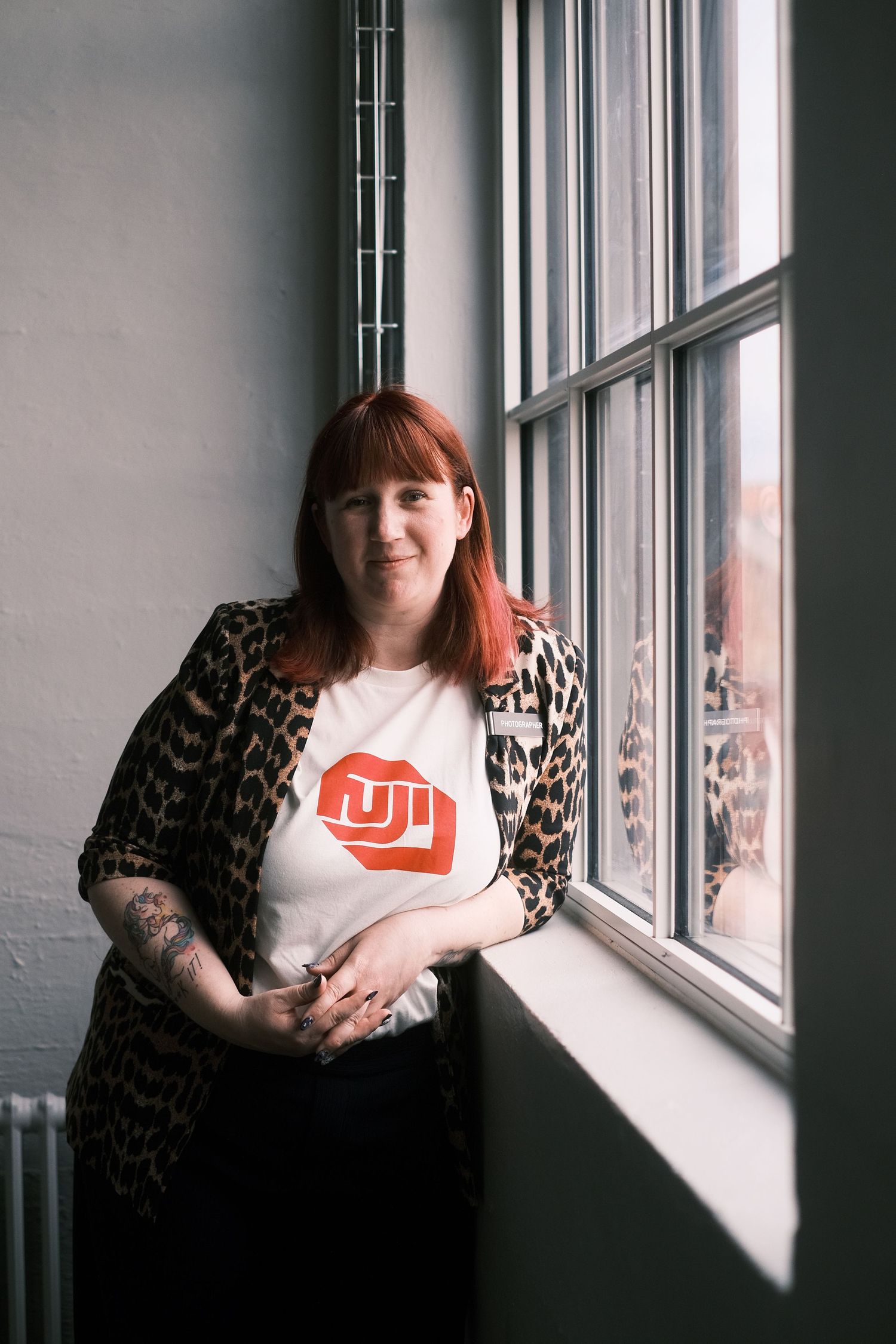 Red-haired woman in leopard print jacket and Fuji t-shirt standing by a window in natural light.