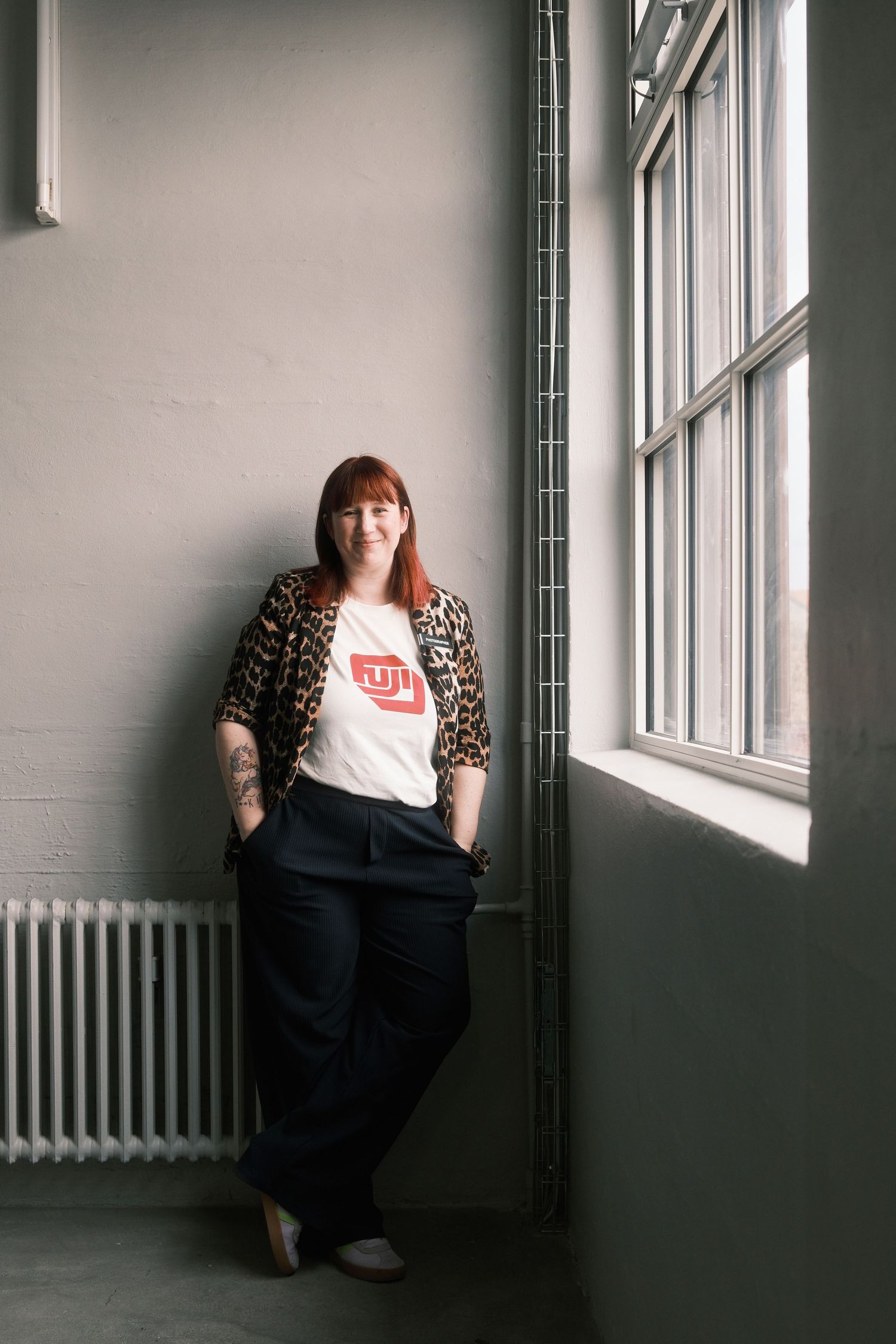 Woman in leopard print jacket and Fuji t-shirt leaning against wall near window.