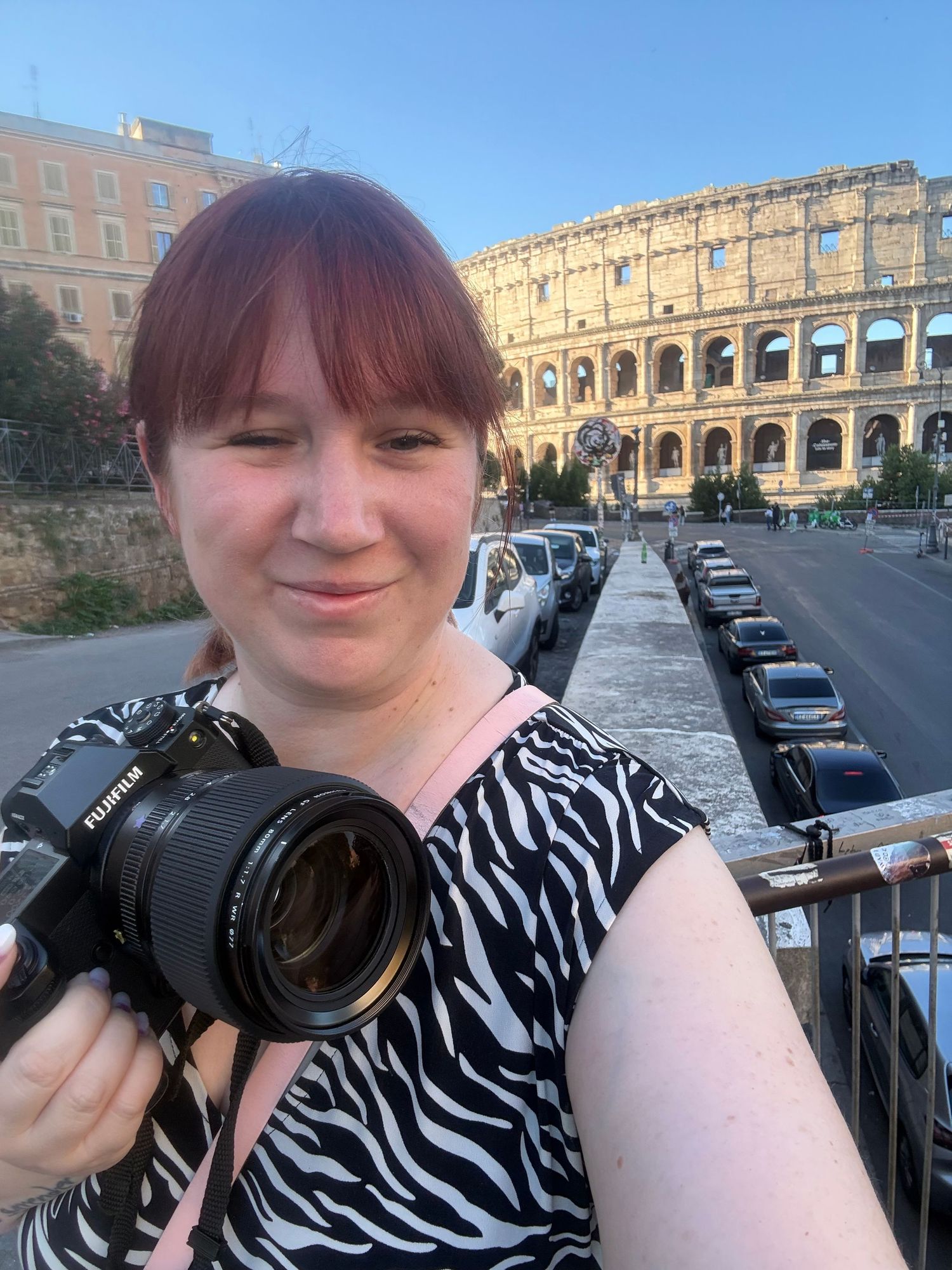 Young woman with red hair holding a Fujifilm camera, selfie in front of the Colosseum in Rome.