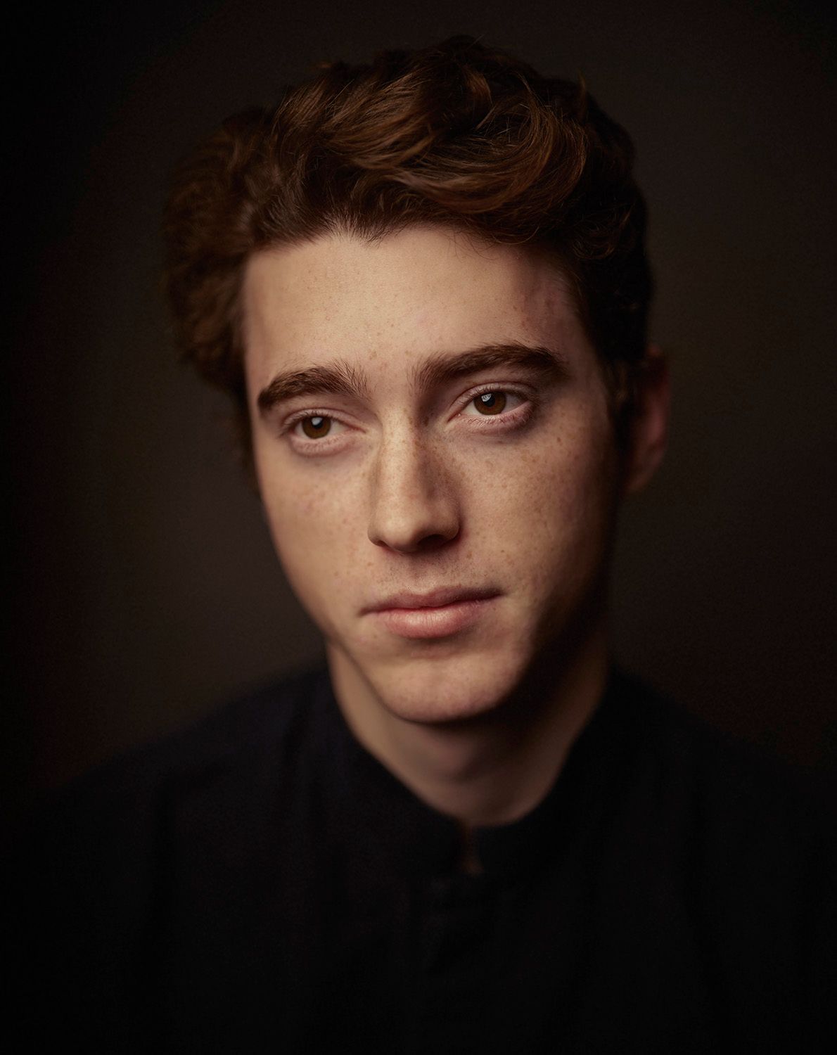 Portrait of a young man with curly red hair and freckles against a dark background.