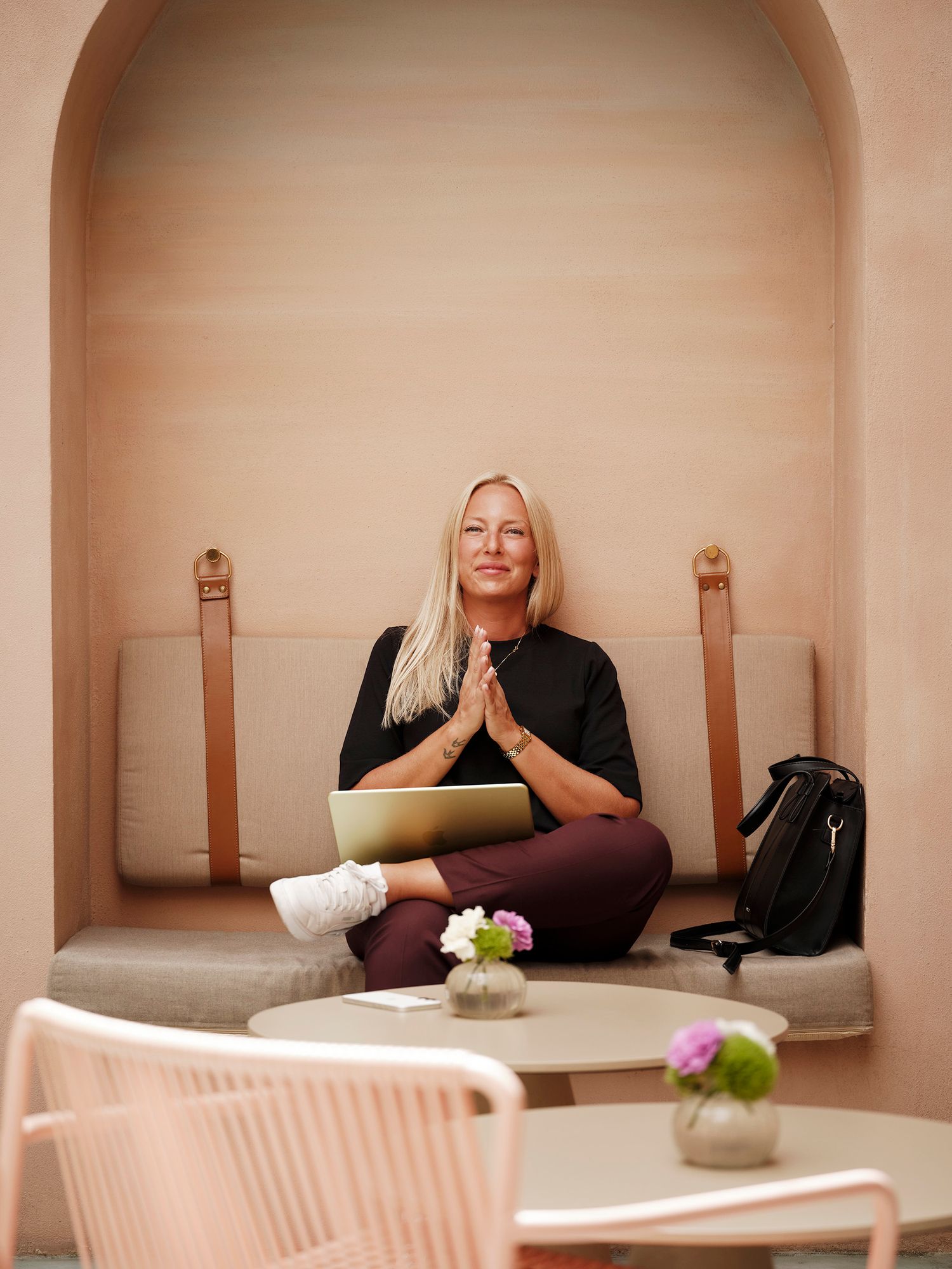 Blonde woman sitting cross-legged on a bench in prayer pose with a laptop, in a pink minimalist alcove.