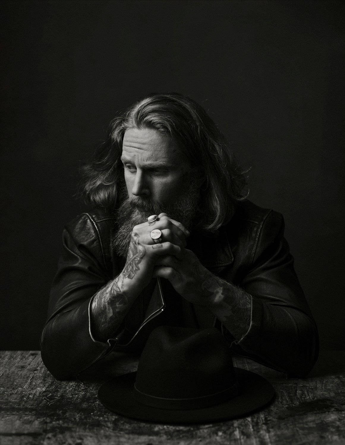 Black and white portrait of a bearded man with long hair in leather jacket, hands clasped over a hat.