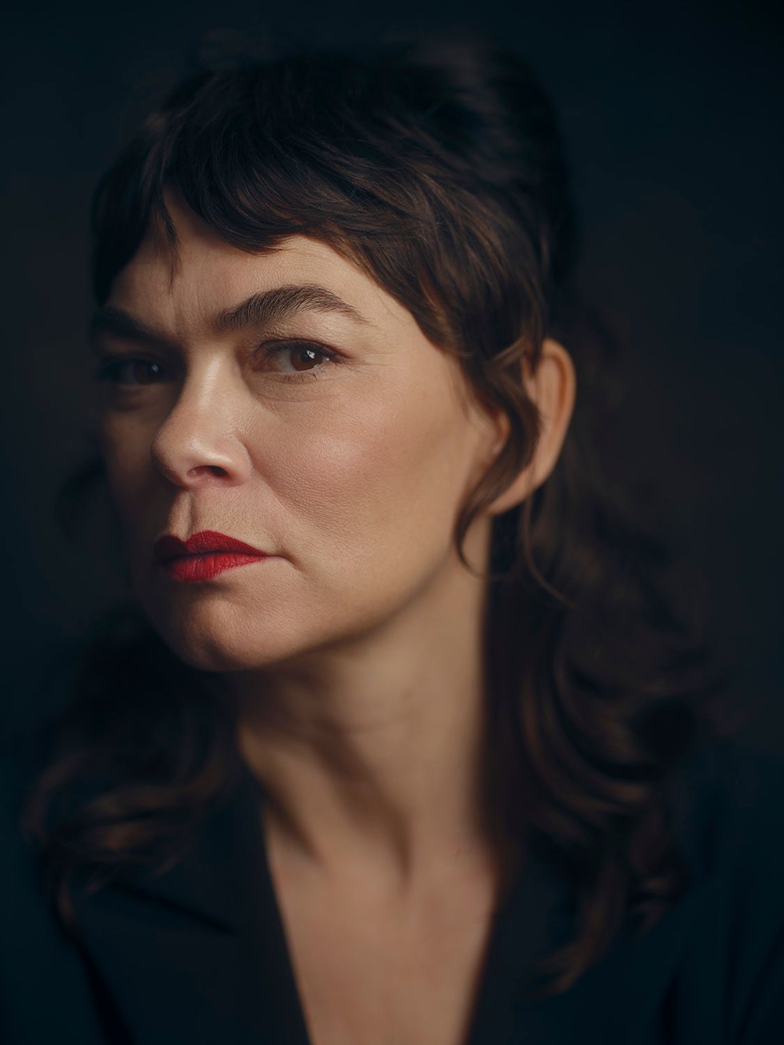 Close-up portrait of a woman with dark wavy hair and red lips against a dark background.