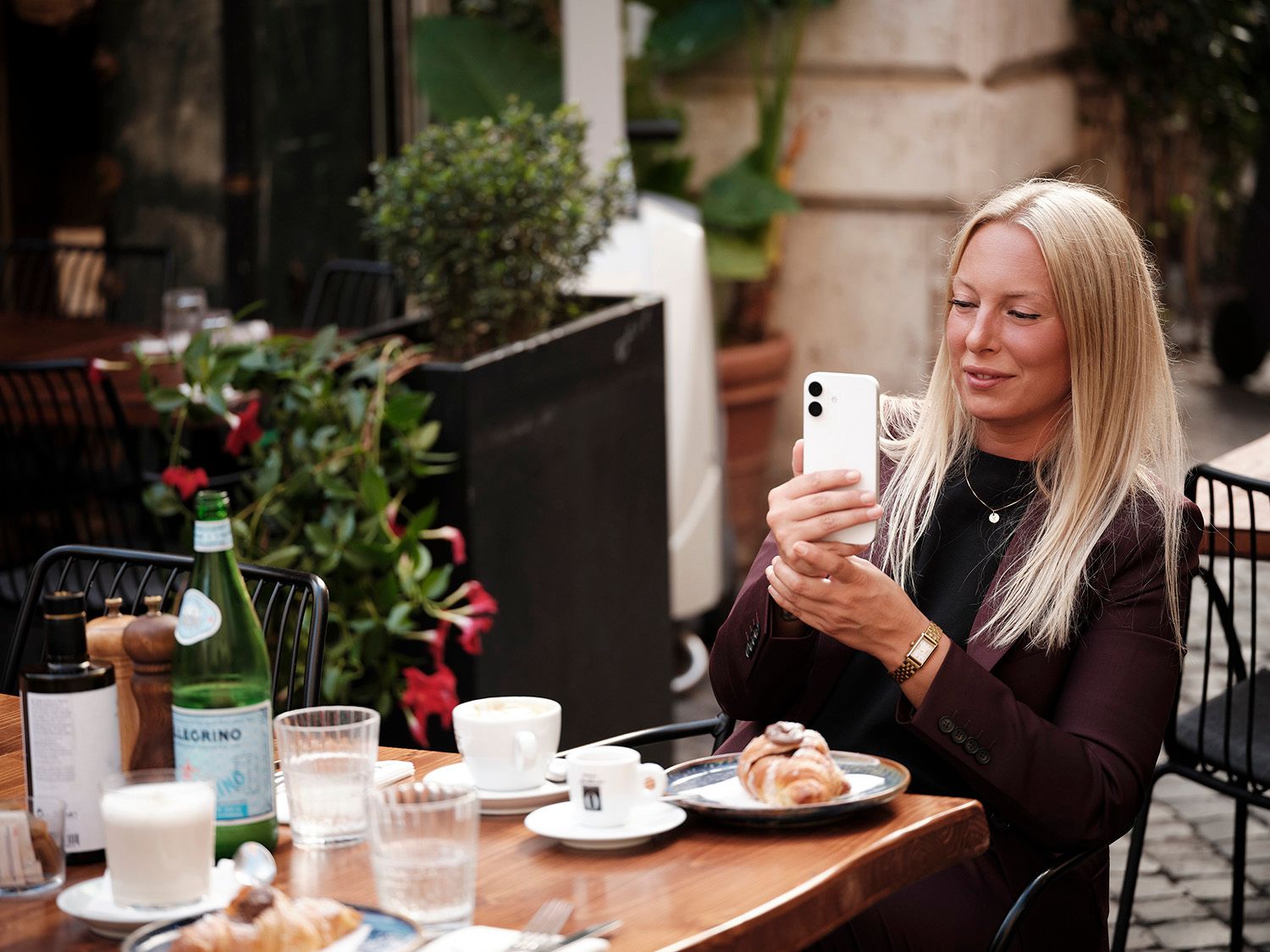 Blonde woman photographing food at an outdoor cafe table in Rome with coffee and croissants.