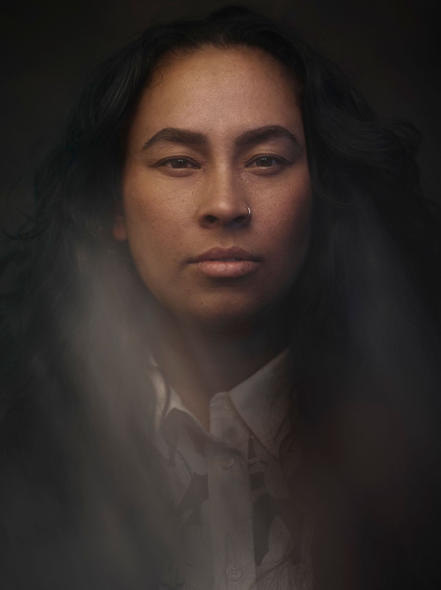 Moody portrait of a woman with dark wavy hair, nose ring, and white shirt against dark background.