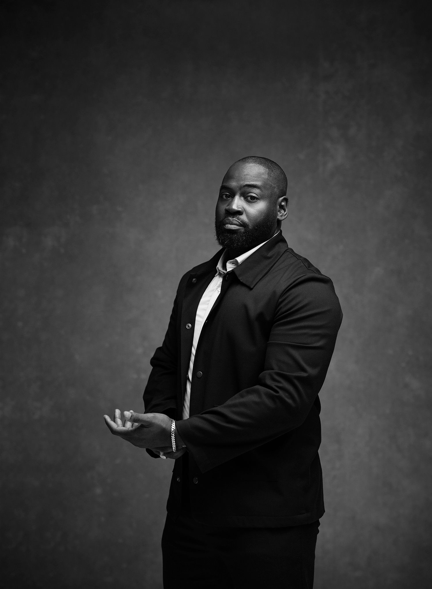Black and white portrait of a man in a dark suit standing against a textured grey background.