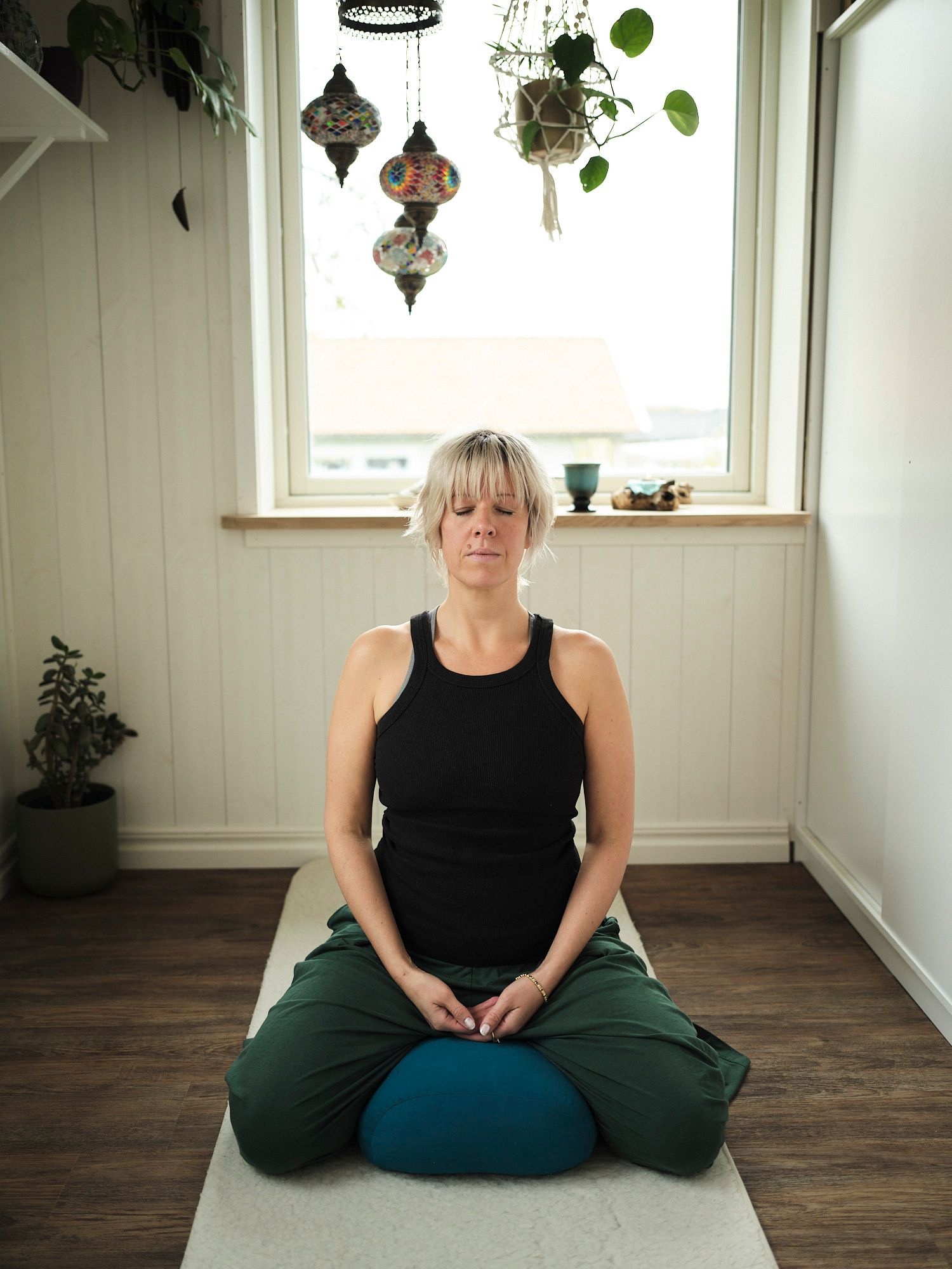 Woman meditating in lotus pose on a yoga mat in a serene room with hanging lanterns and plants.