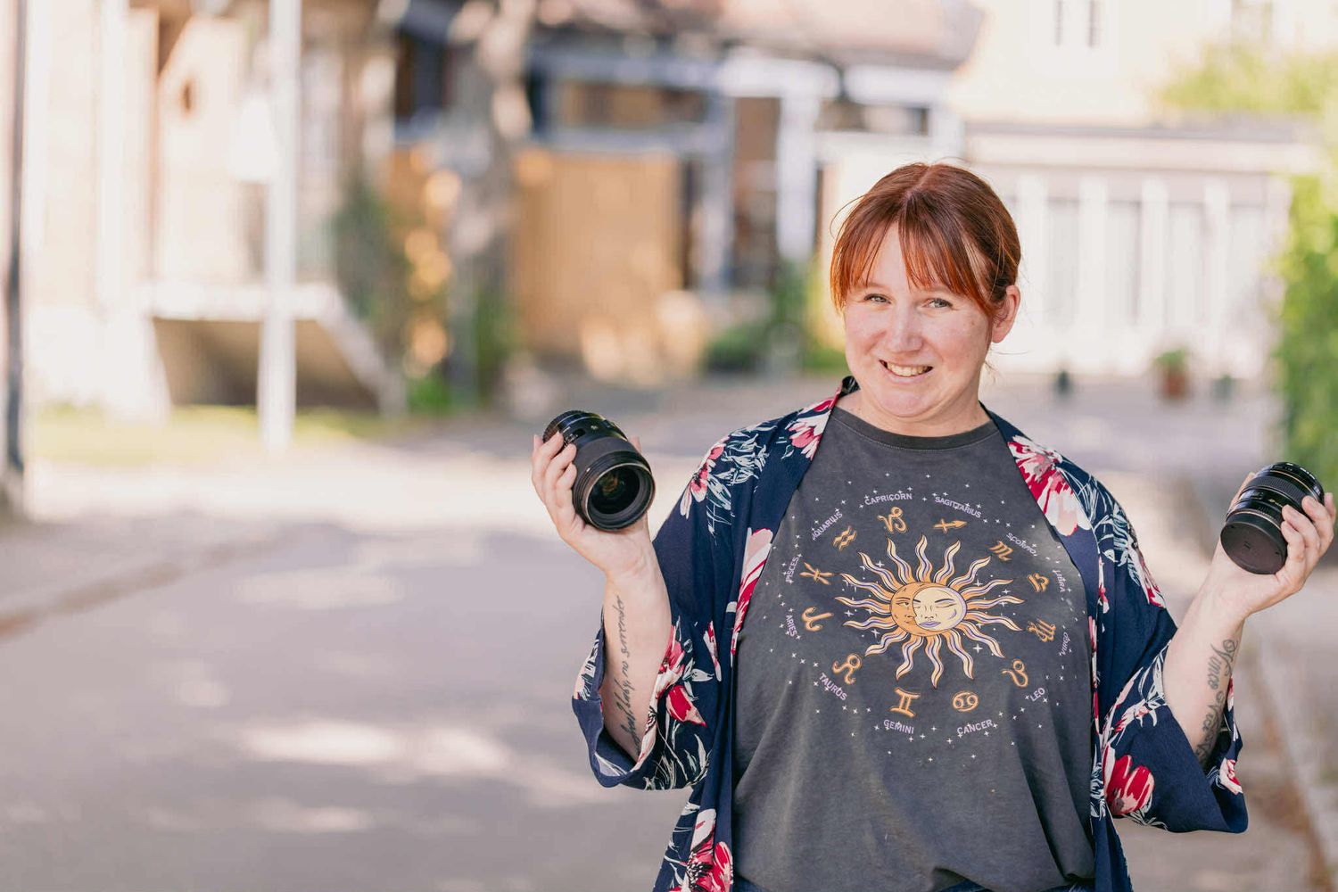 Smiling woman holding two cameras outdoors on sunny street.