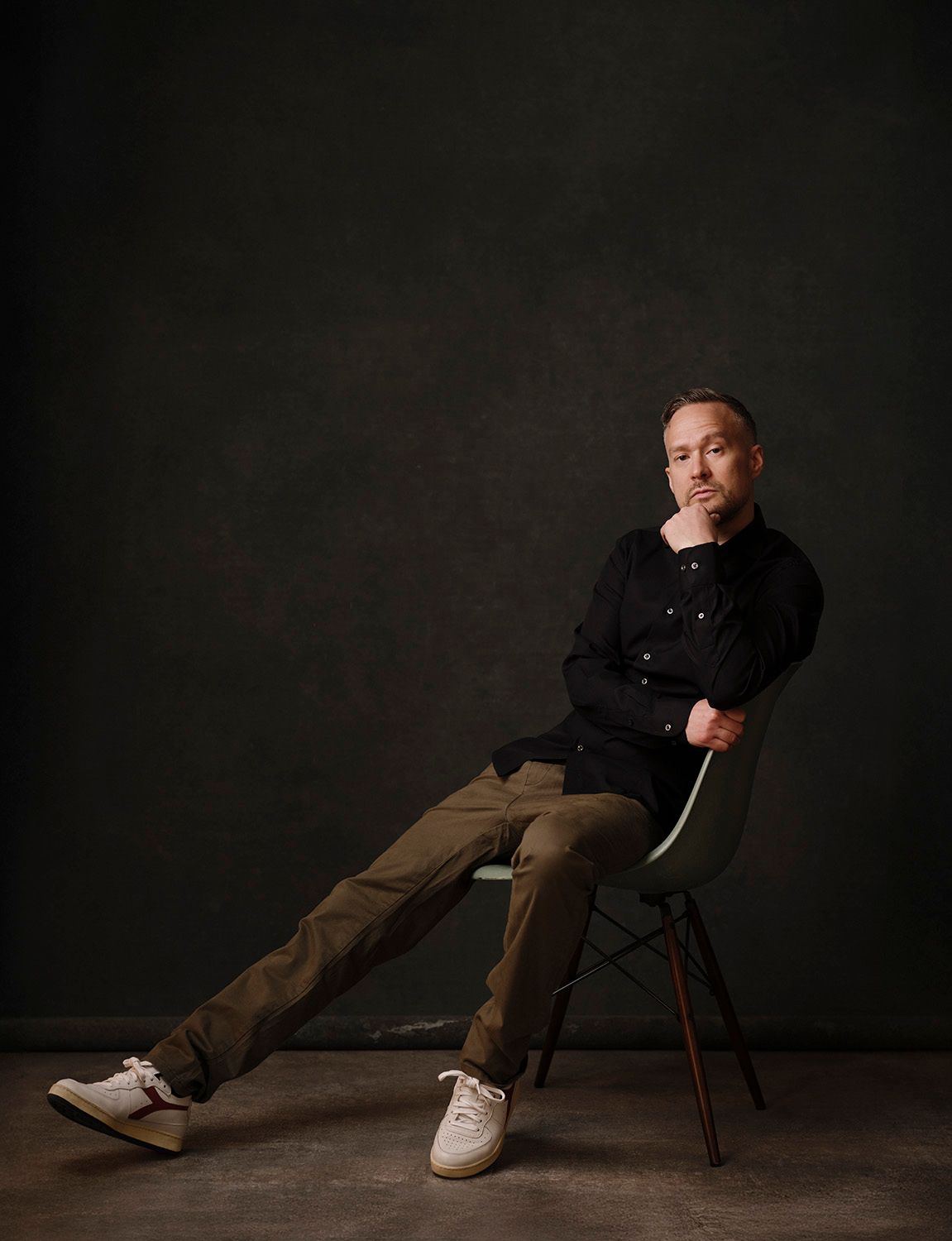 Man in black jacket and khaki pants sitting casually on a chair against a dark green background.