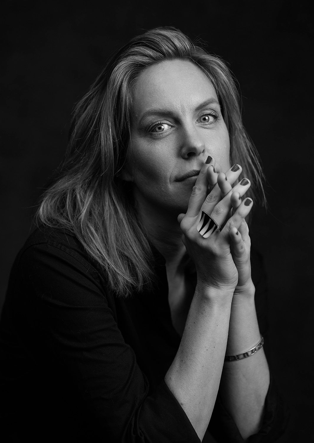 Black and white portrait of a woman with long hair, hands clasped near her face, on dark background.