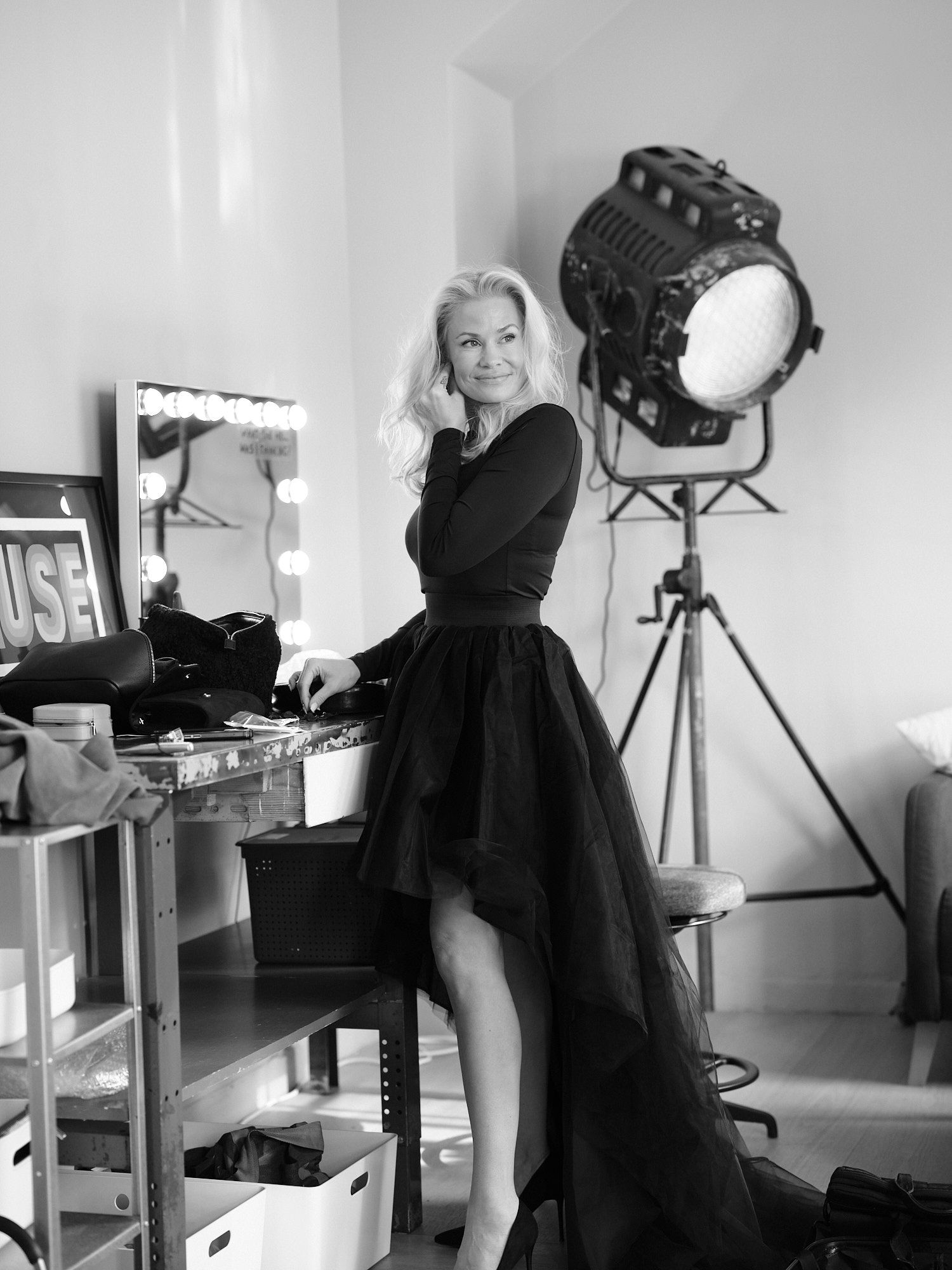 Black and white photo of a blonde woman in black asymmetric dress posing in a photography studio.
