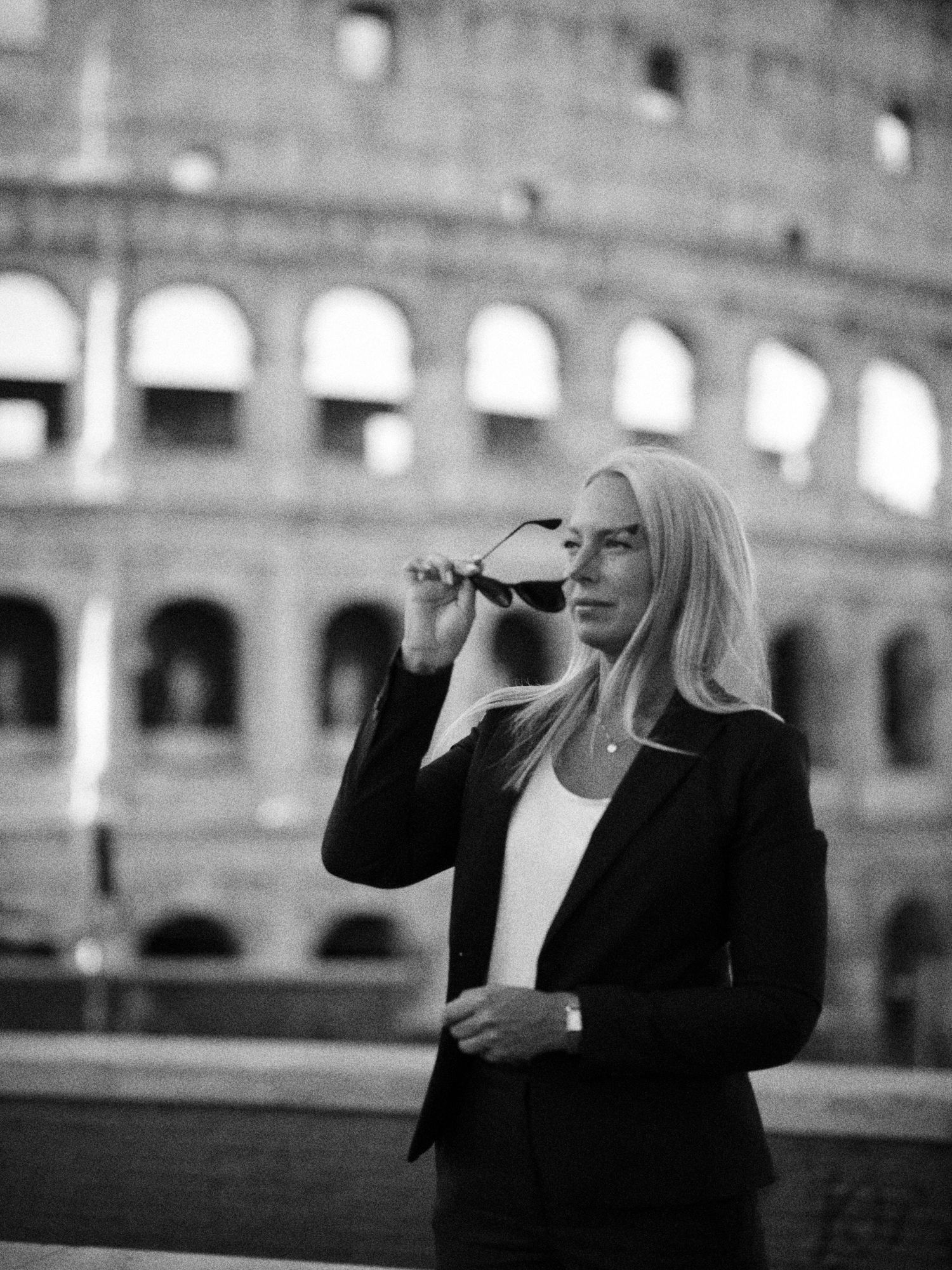 Elegant blonde woman holding sunglasses in front of the Colosseum in Rome, black and white photo.