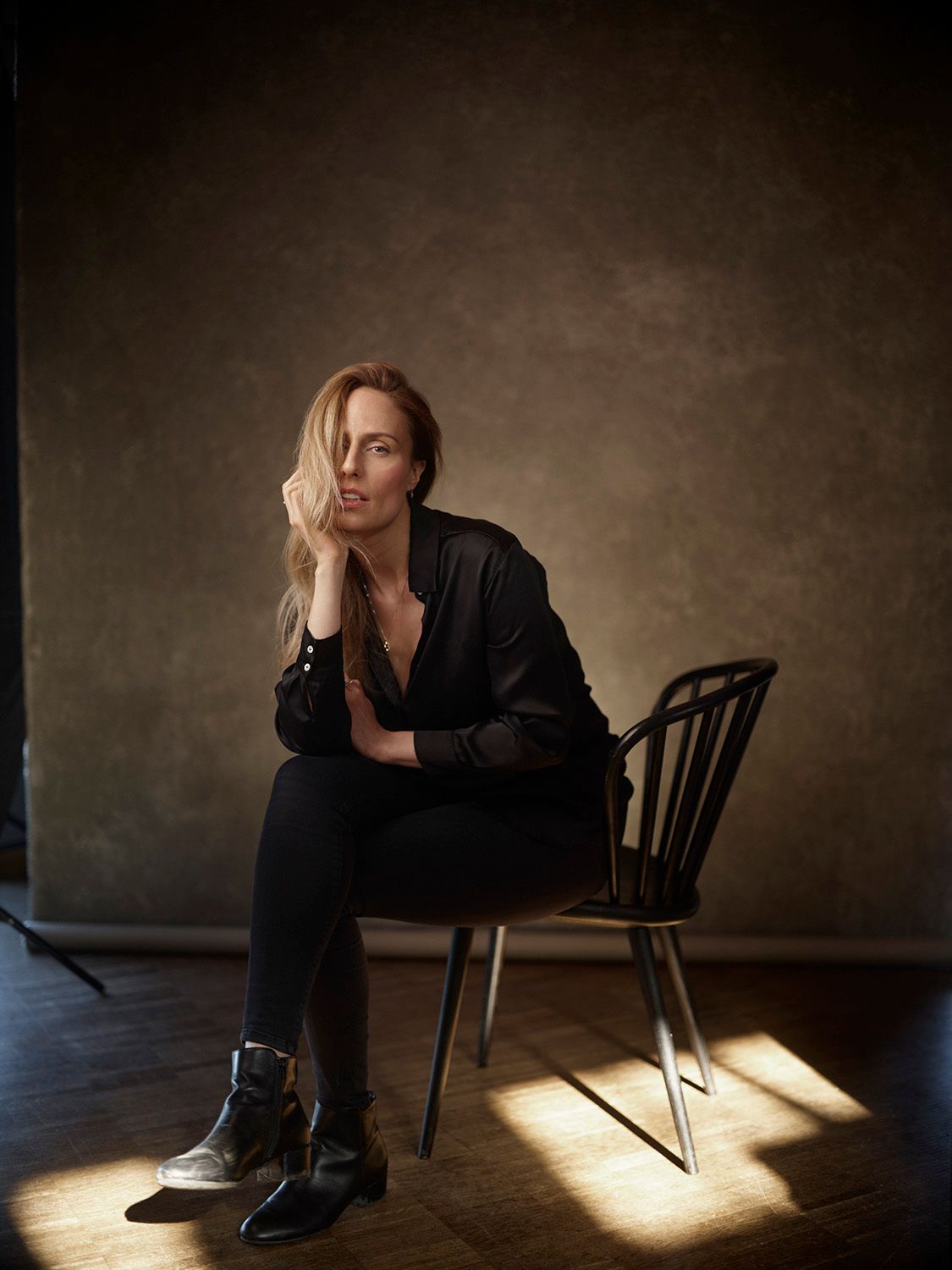 Blonde woman in black outfit seated in chair with dramatic lighting.