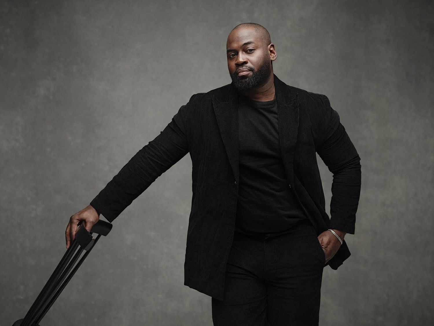 Confident Black man in all-black suit leaning on a cane against a neutral gray studio background.