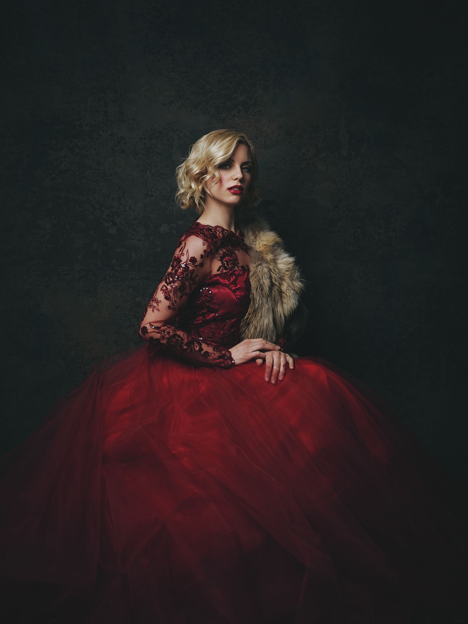Elegant woman in a stunning red ballgown with lace sleeves and fur stole against dark background.