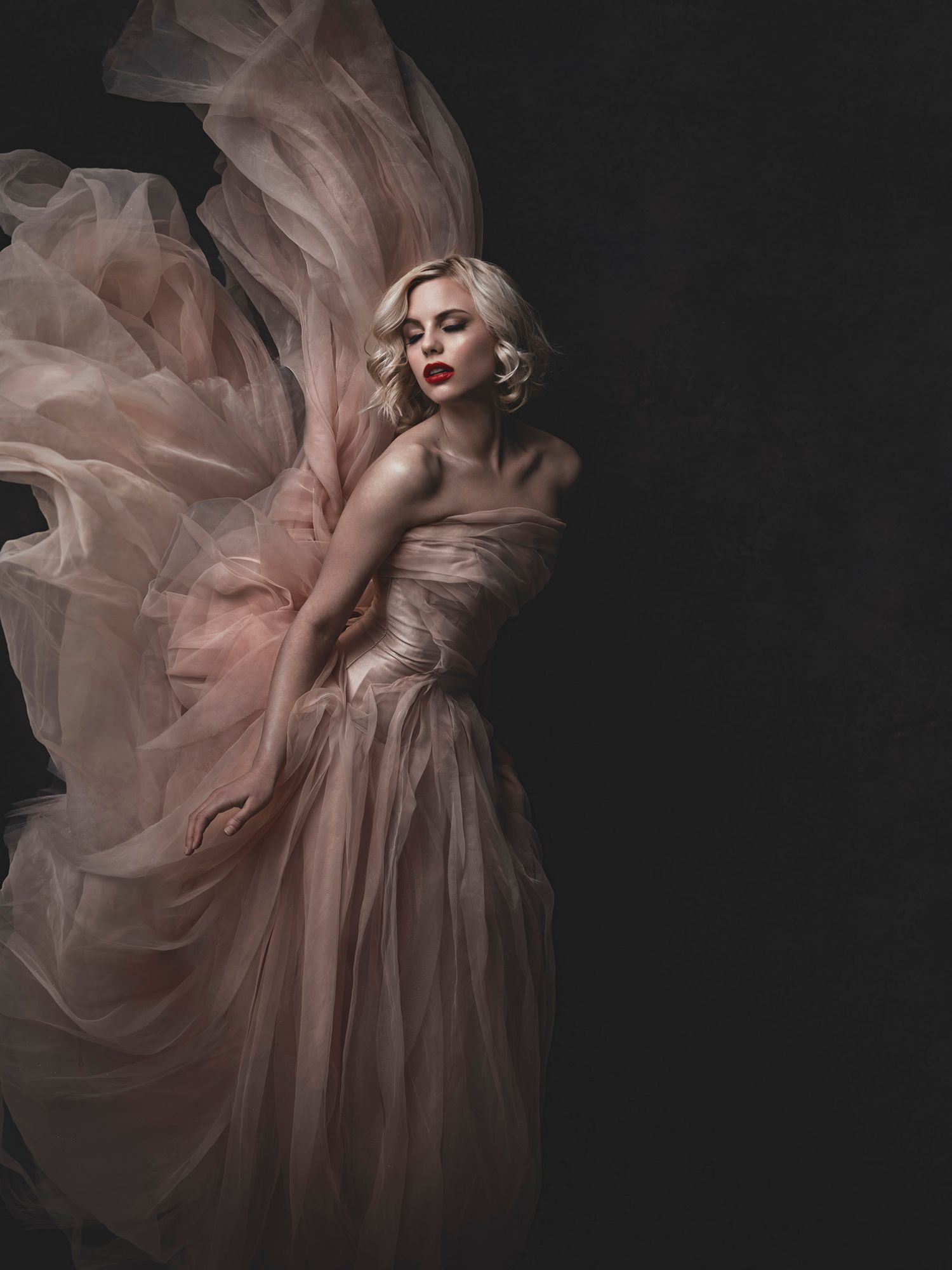 Blonde woman in a flowing blush pink tulle gown posed dramatically against a dark background.