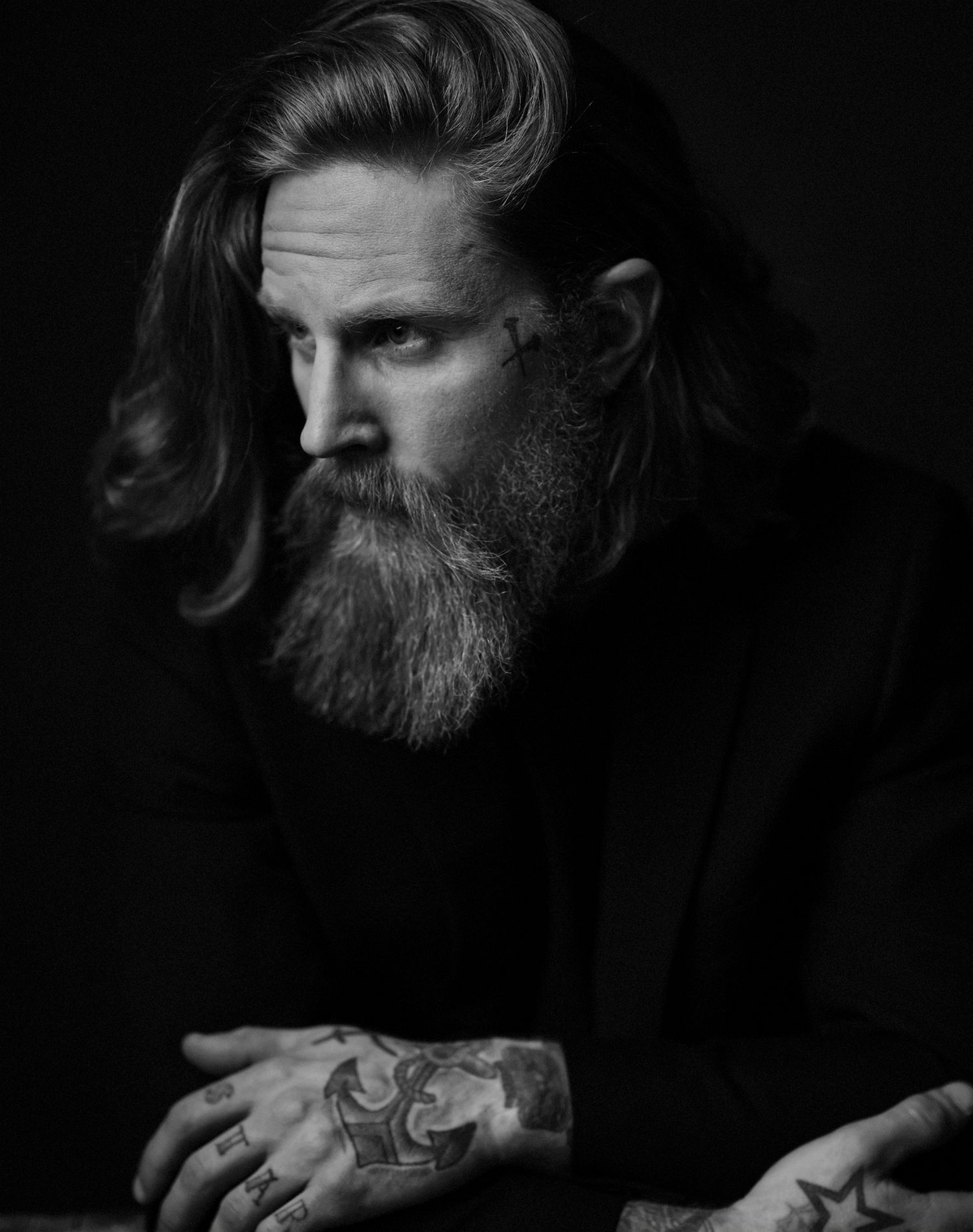 Black and white portrait of a bearded man with long hair, tattoos on hands, wearing a black top.