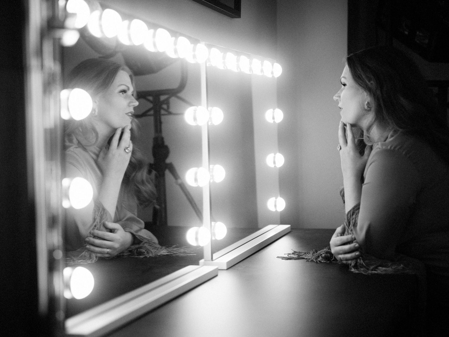 Black and white photo of a woman applying makeup at a vanity mirror with Hollywood lights.