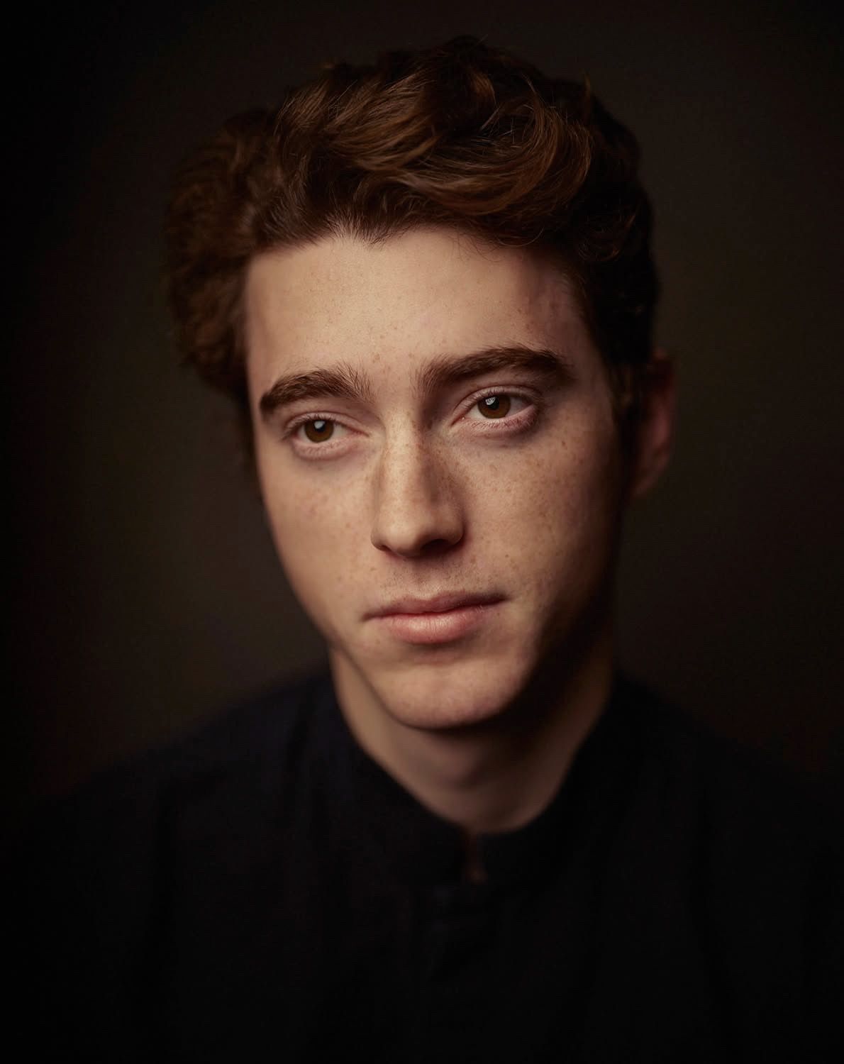 Young man with auburn curly hair and freckles in dark dramatic portrait.