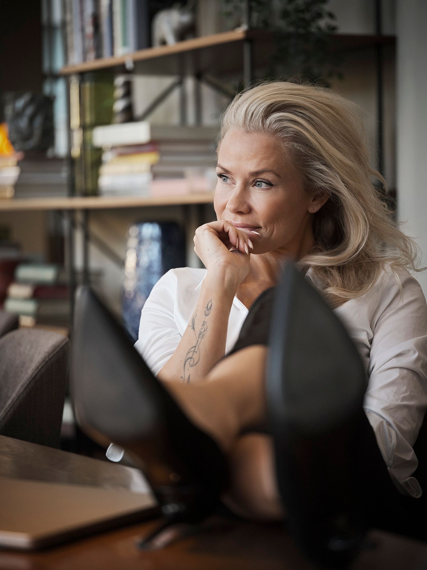 Mature blonde woman with tattoo resting chin on hand, gazing thoughtfully in a modern office setting.