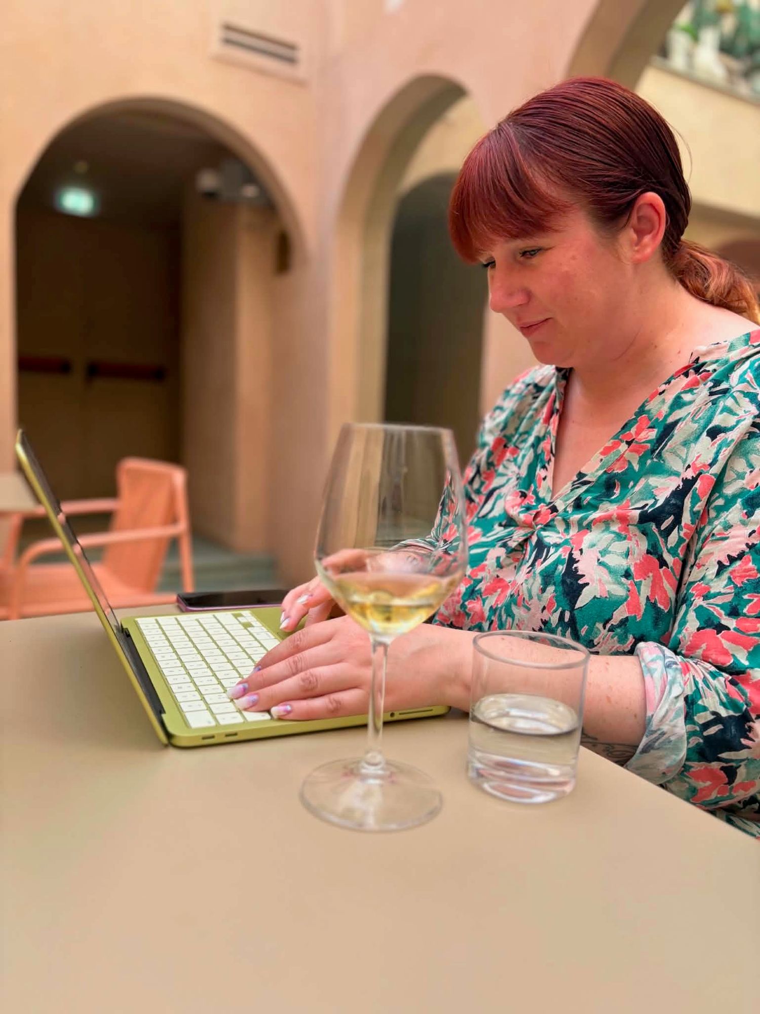 Woman working on laptop with white wine glass at outdoor cafe table.