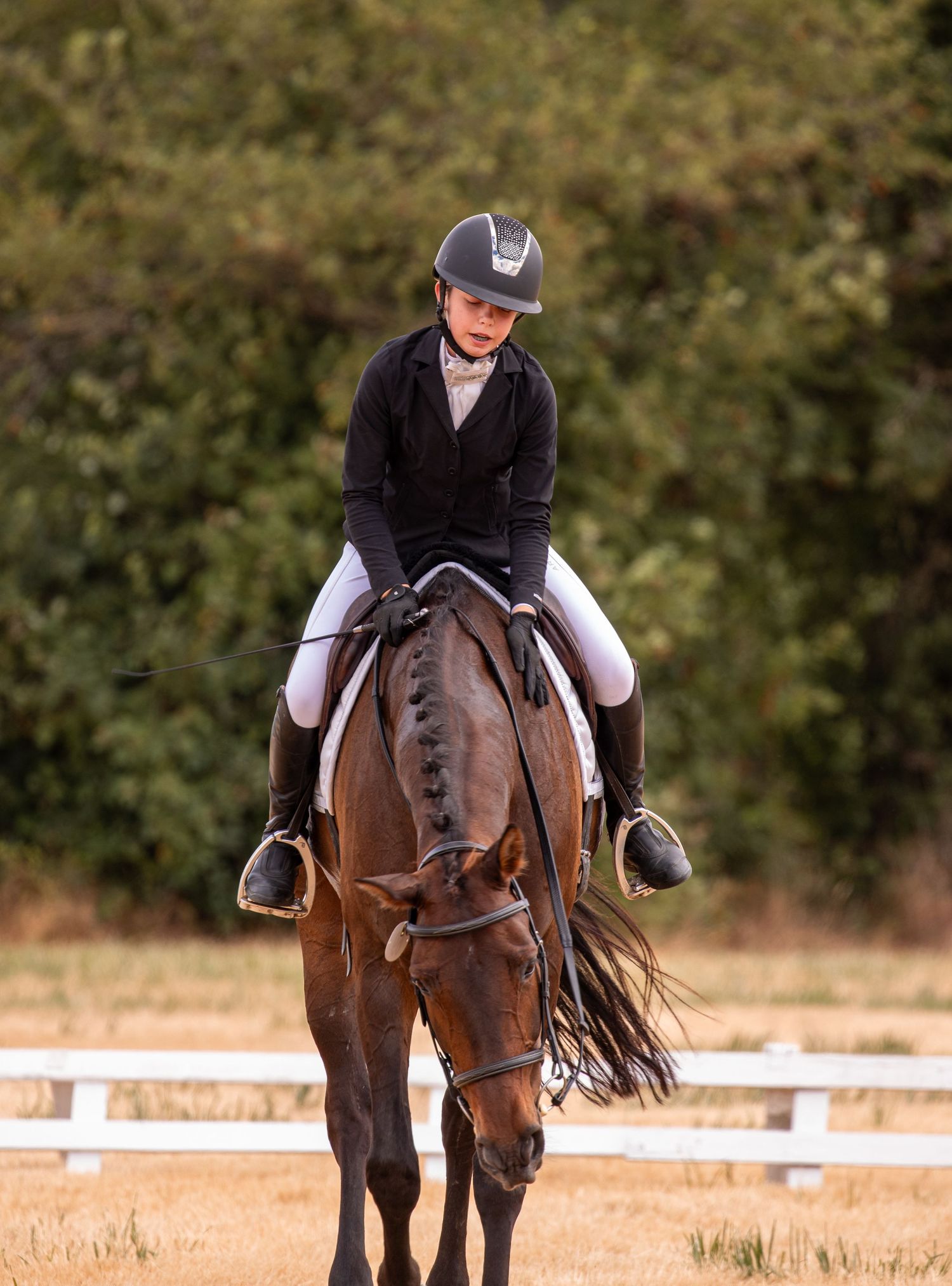 Female rider in black dressage attire riding a bay horse in a white-fenced outdoor arena.