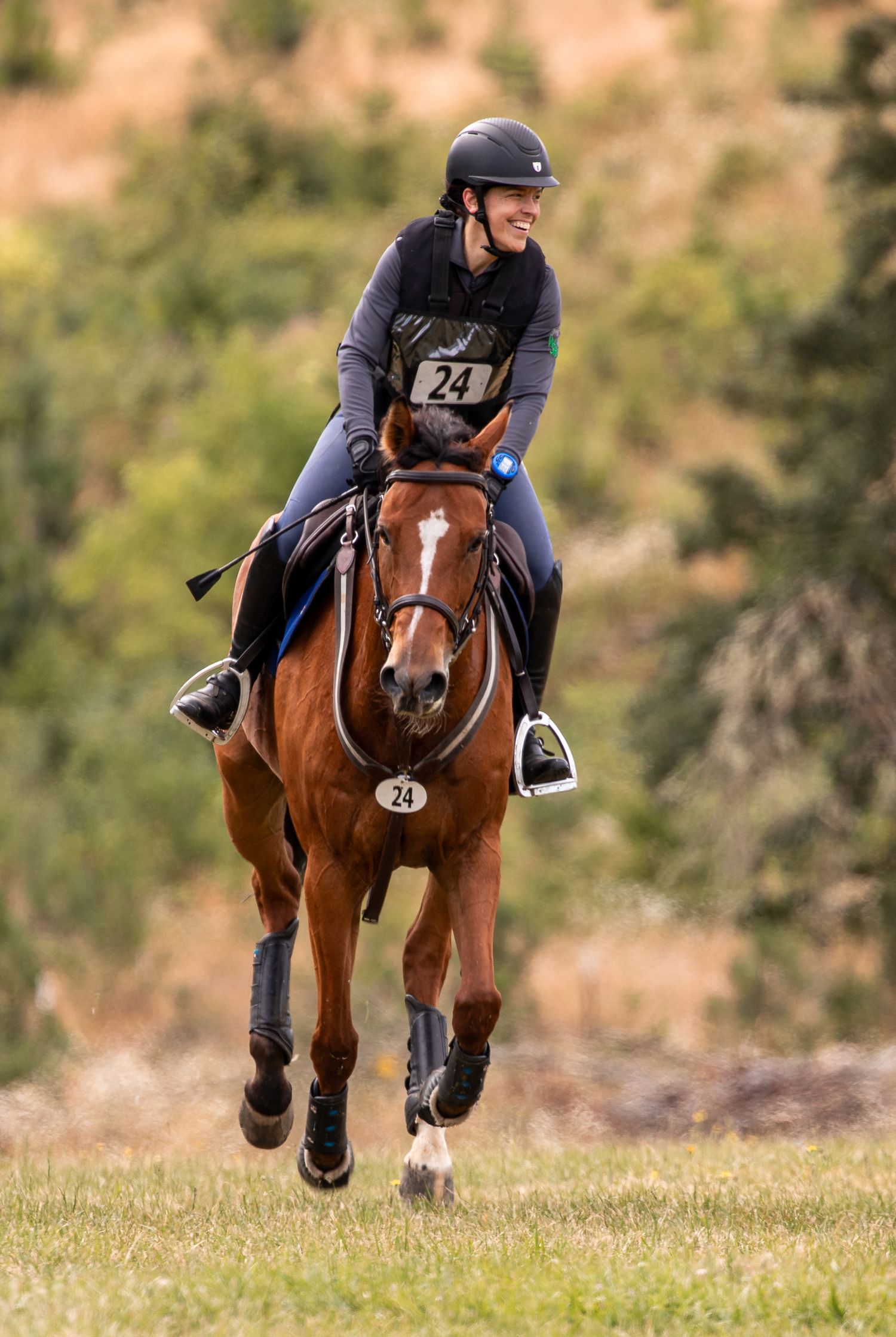 Smiling rider wearing number 24 galloping a chestnut horse across a green cross-country course.