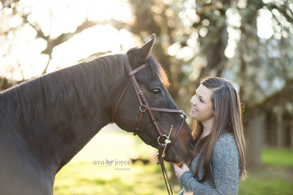 A girl poses with her horse in a field with the sun shining through the trees.