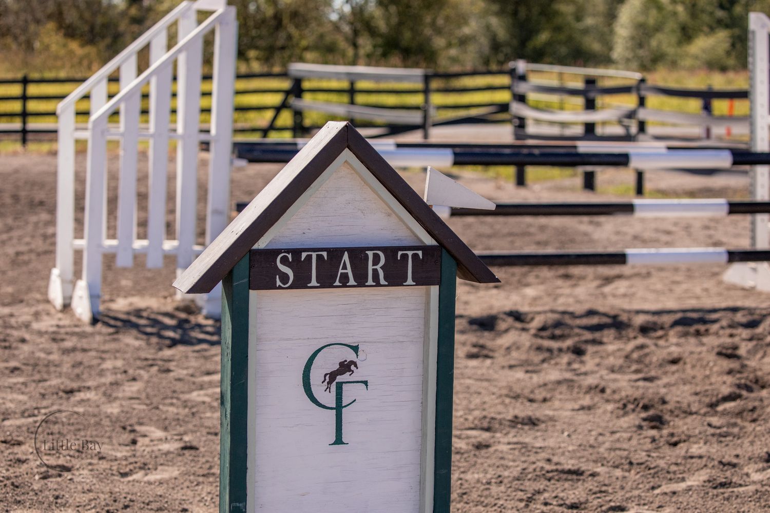 A START marker sign with a green logo stands in a sandy equestrian arena with white jump fences in the background.