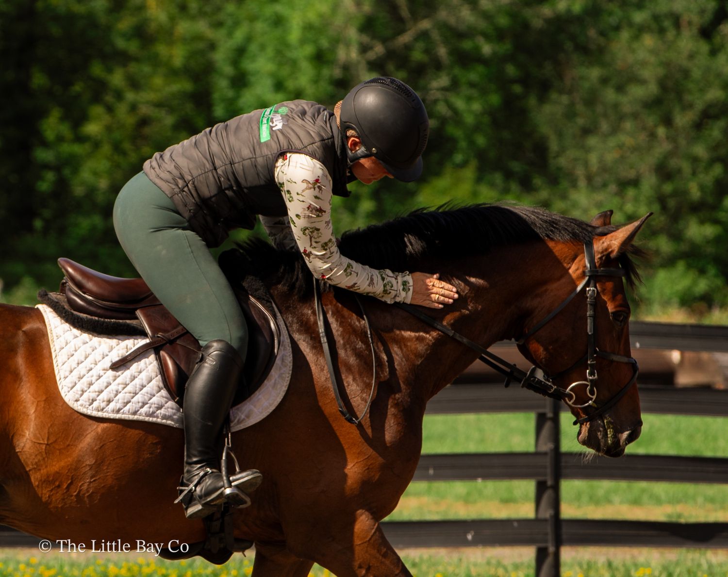 Equestrian rider in black helmet and green breeches leaning forward on a bay horse at an outdoor riding arena.