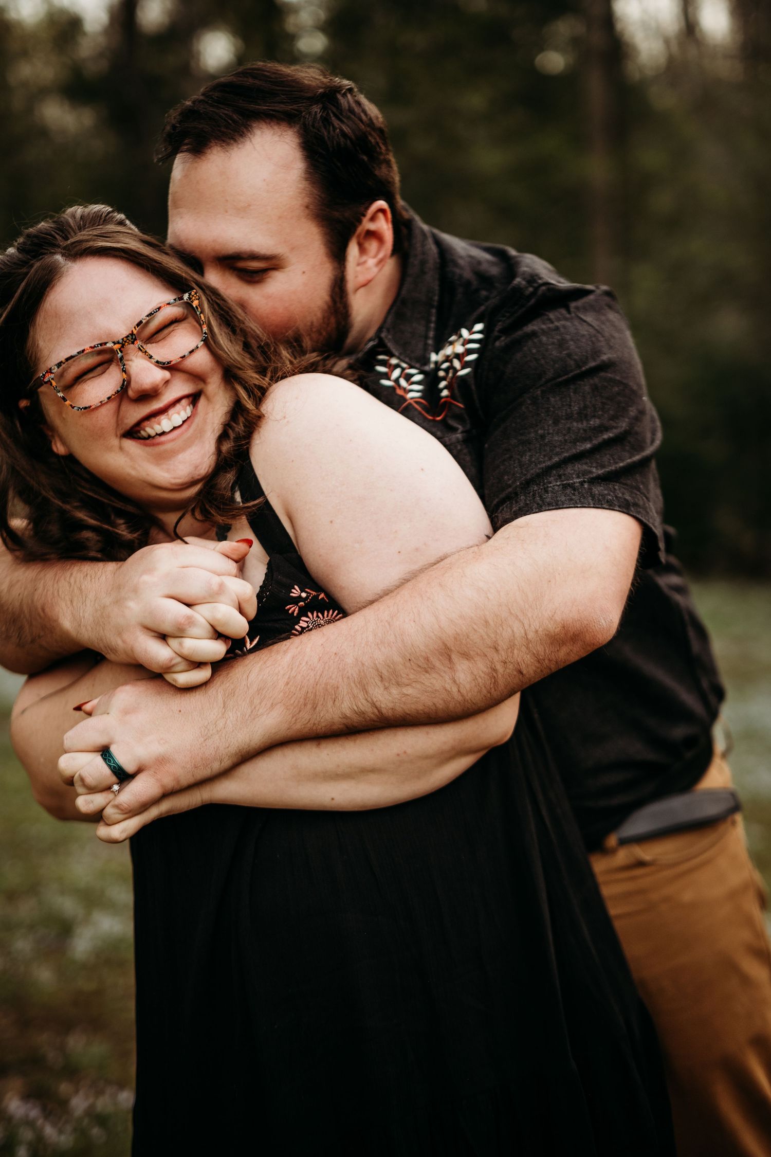 Man wrapping a woman up in a bear hug in his arms, kissing her temple as she grins. Engagement session at Audra River State Park in WV