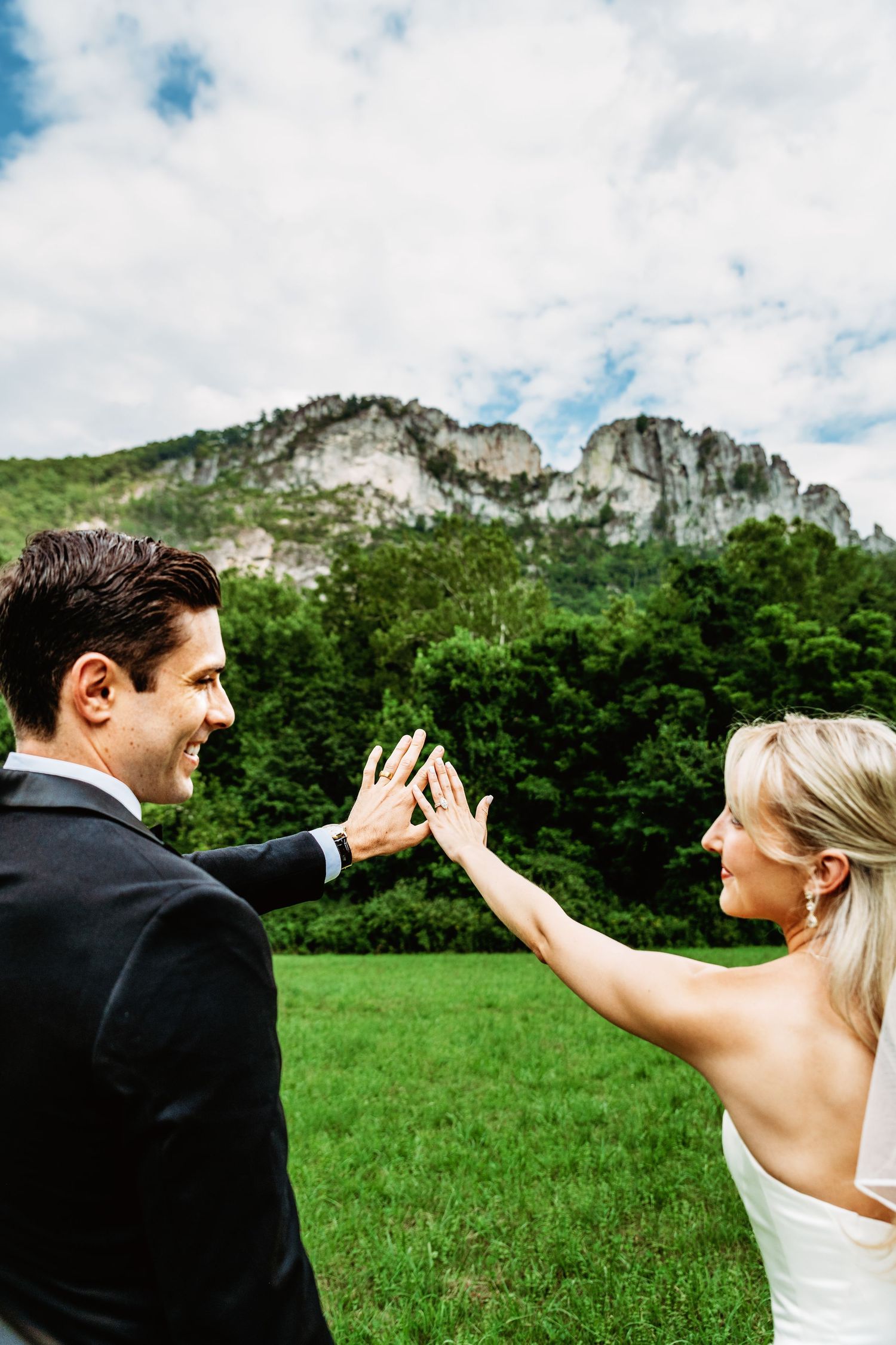 Bride and groom share a smile as they pose with their rings up in the air in front of them, showing off Seneca Rocks, WV in the background