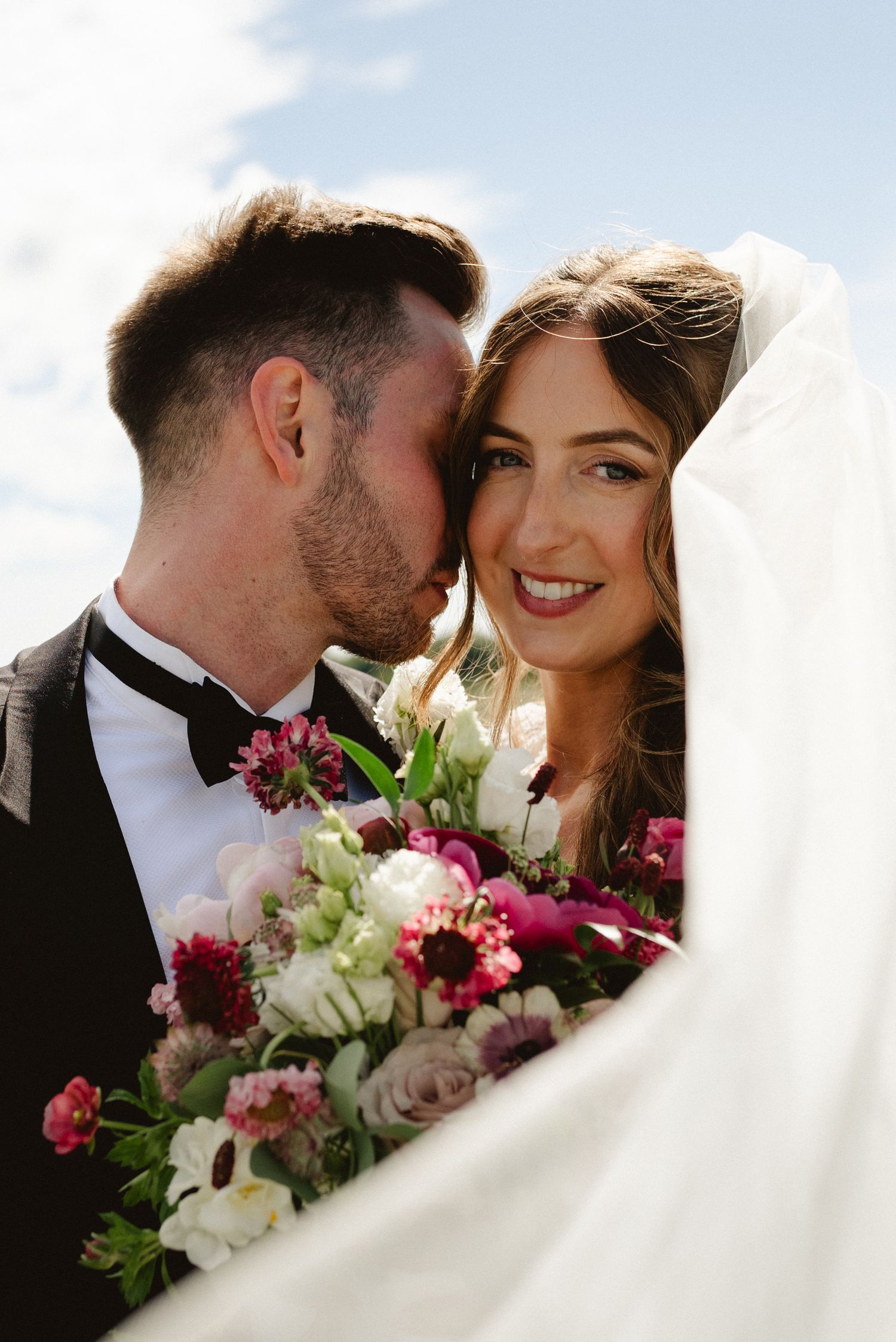 Groom kissing bride on cheek as she smiles holding pink and white floral bouquet under flowing veil.