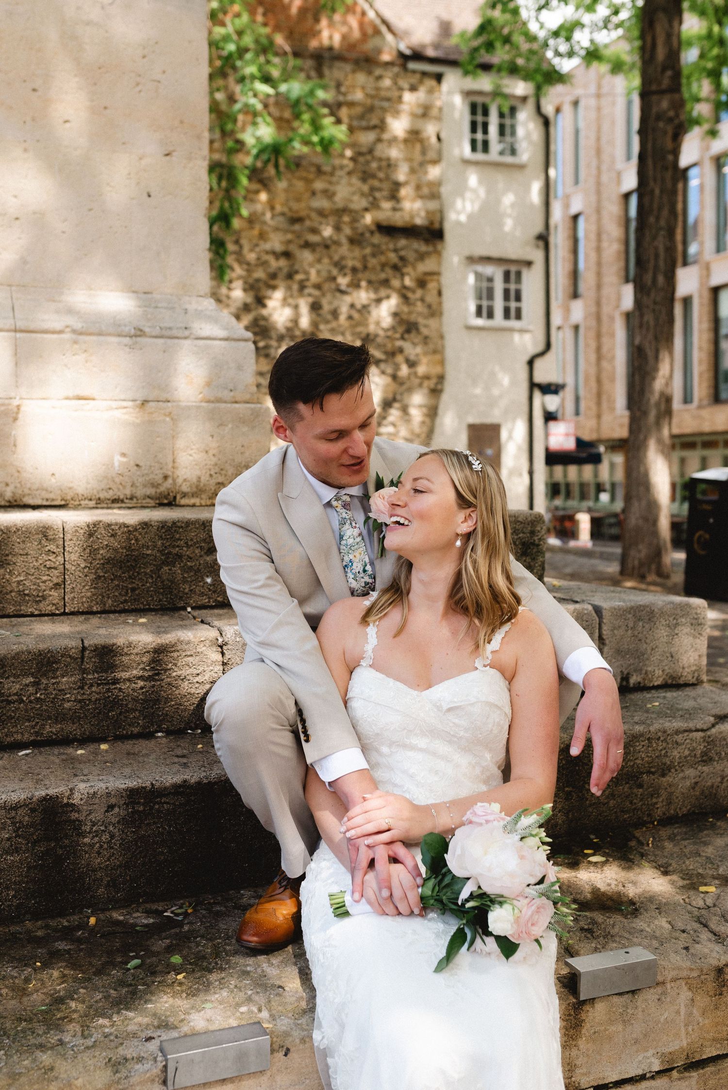 A bride and groom share a joyful moment on stone steps, bride holding white peonies, groom in tan suit.