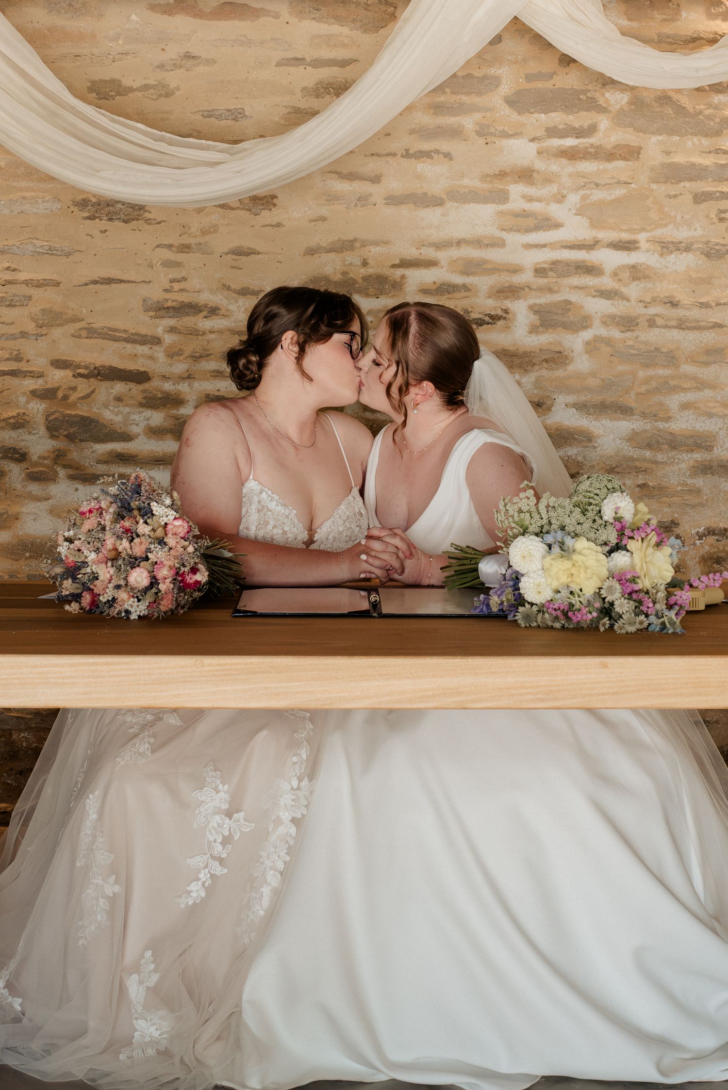 Two brides share a kiss at the signing table, holding hands with floral bouquets beside them in a rustic stone venue.