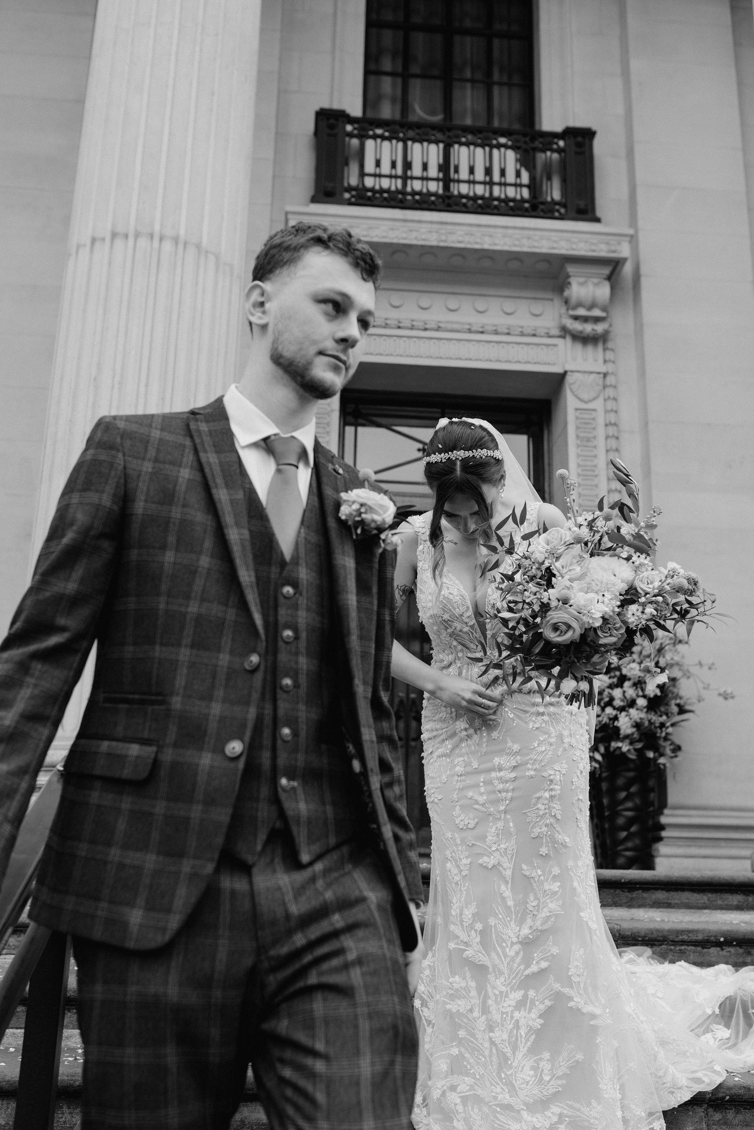 A bride and groom pose outside a classic building with columns, the bride holding a large floral bouquet.