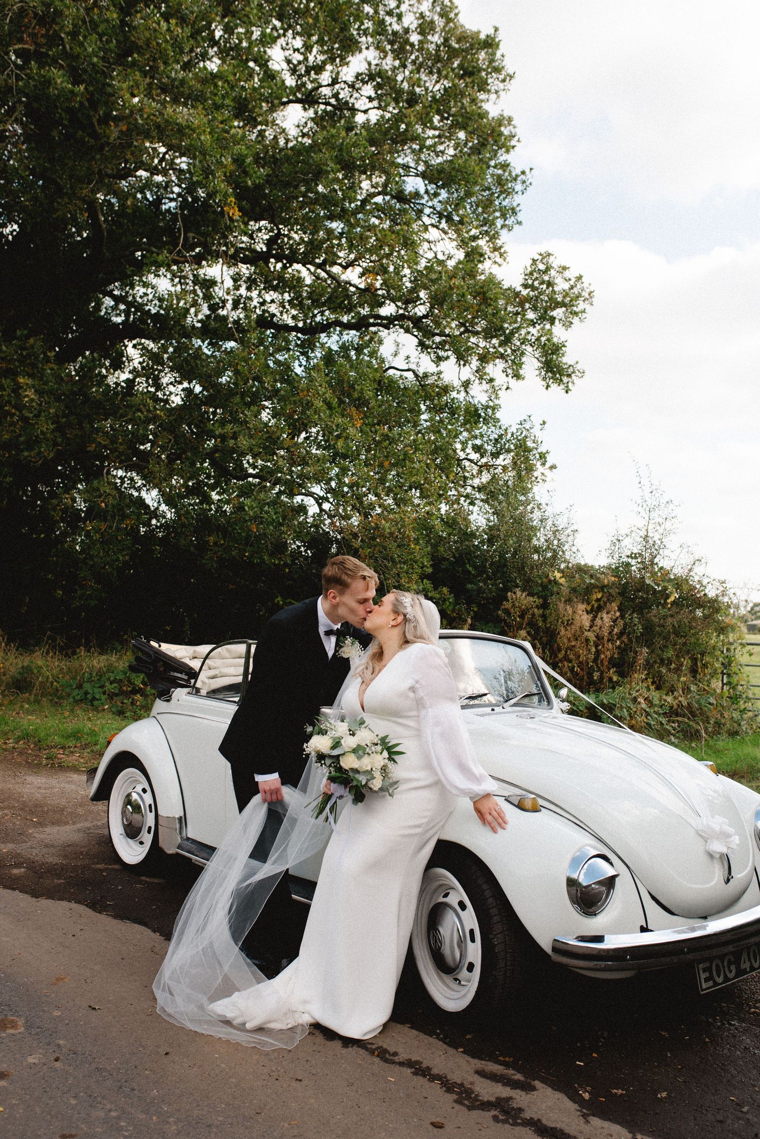 Newlyweds pose together against a classic white Volkswagen Beetle on a country road.