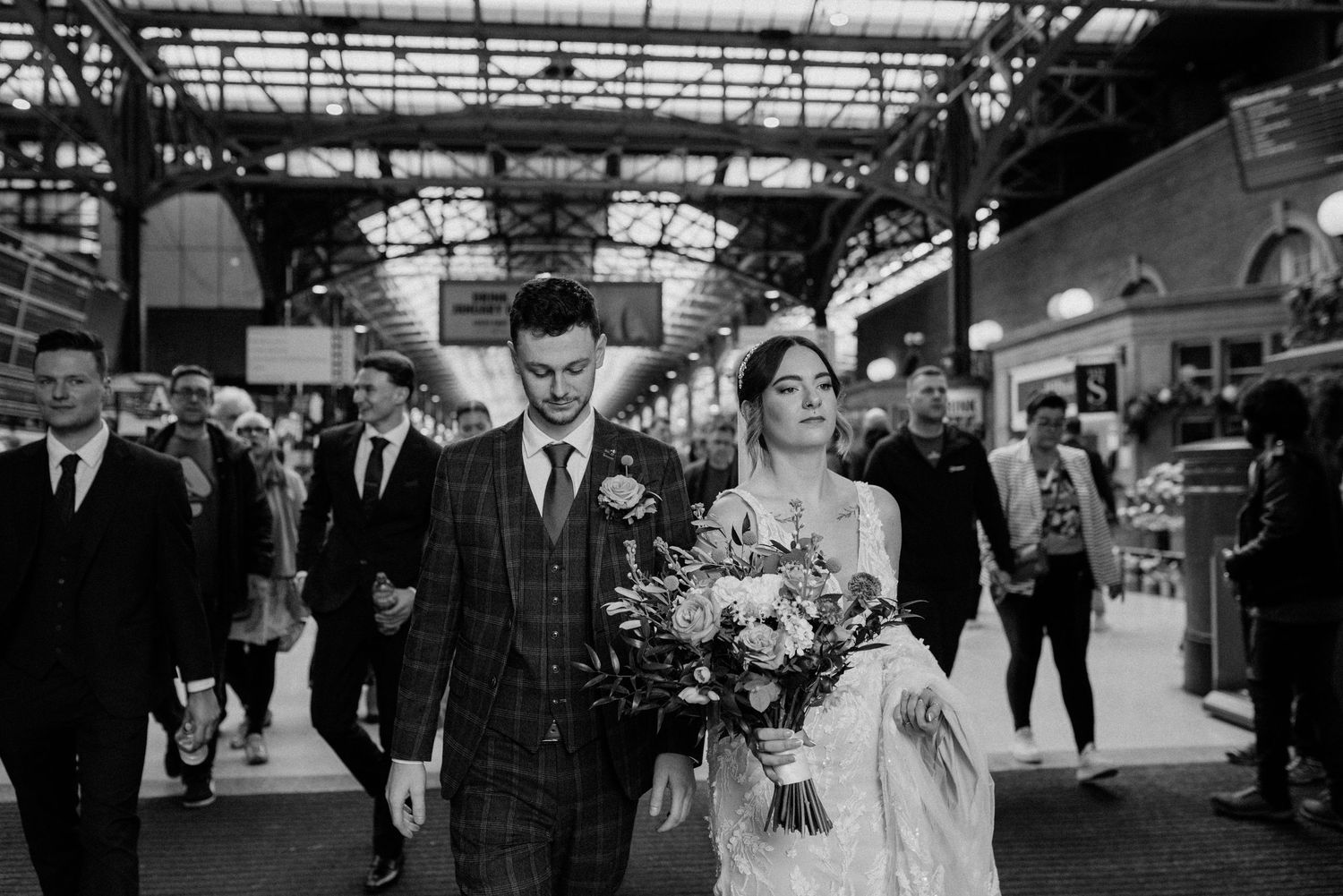 Black and white photo of a bride and groom walking through a busy indoor market, bride holding a floral bouquet.