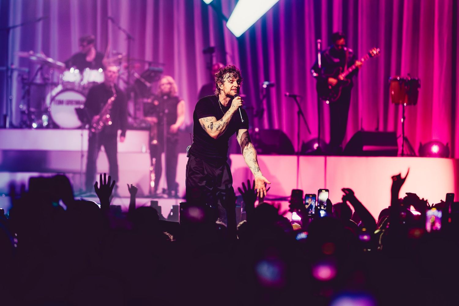 Tom Grennan on stage with purple lighting and crowd silhouettes visible below.