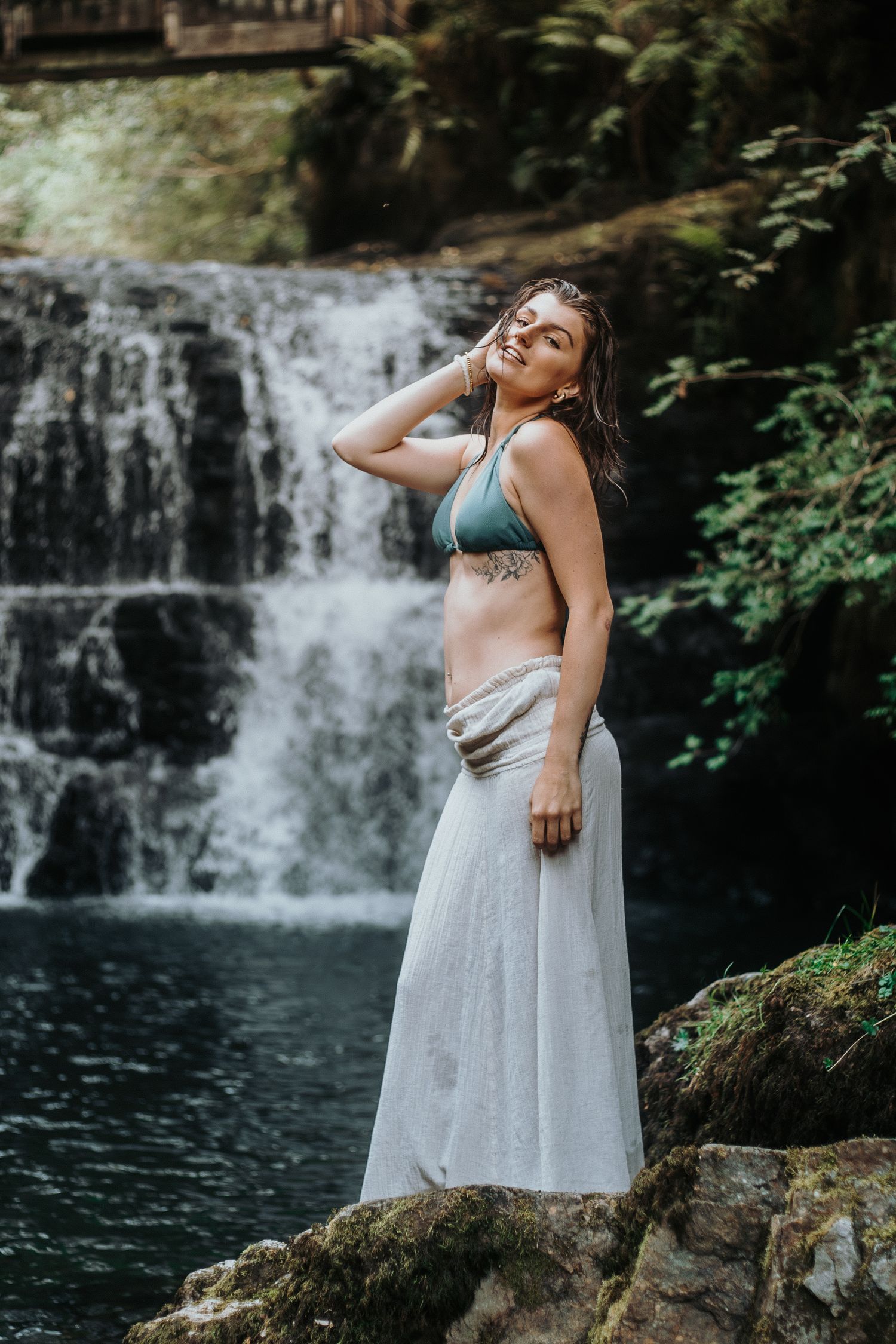 A person in a turquoise bikini top and white skirt poses gracefully on rocks beside a cascading waterfall in nature.