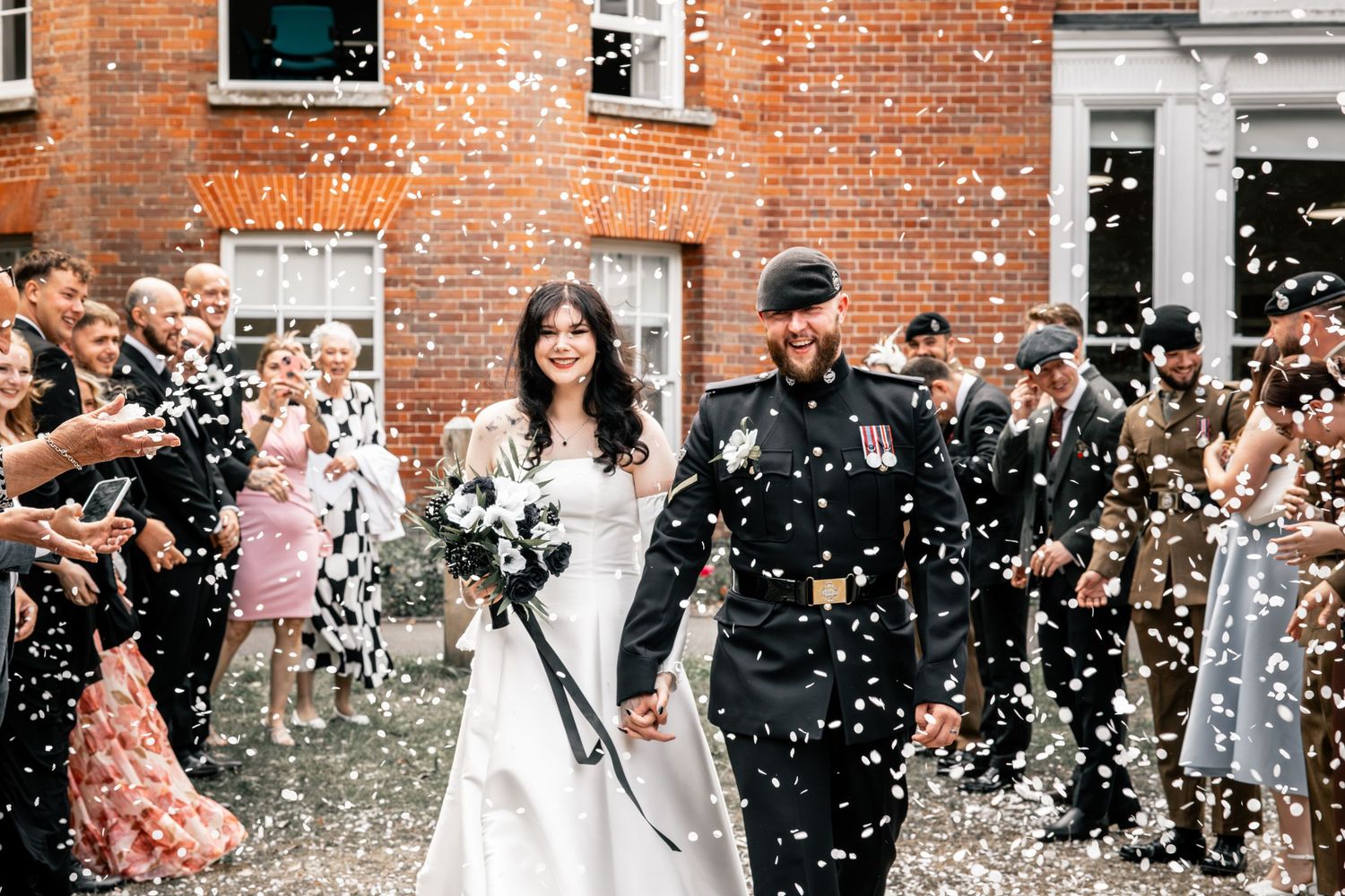 A joyful couple exits their wedding ceremony while guests shower them with white confetti outside a brick building.